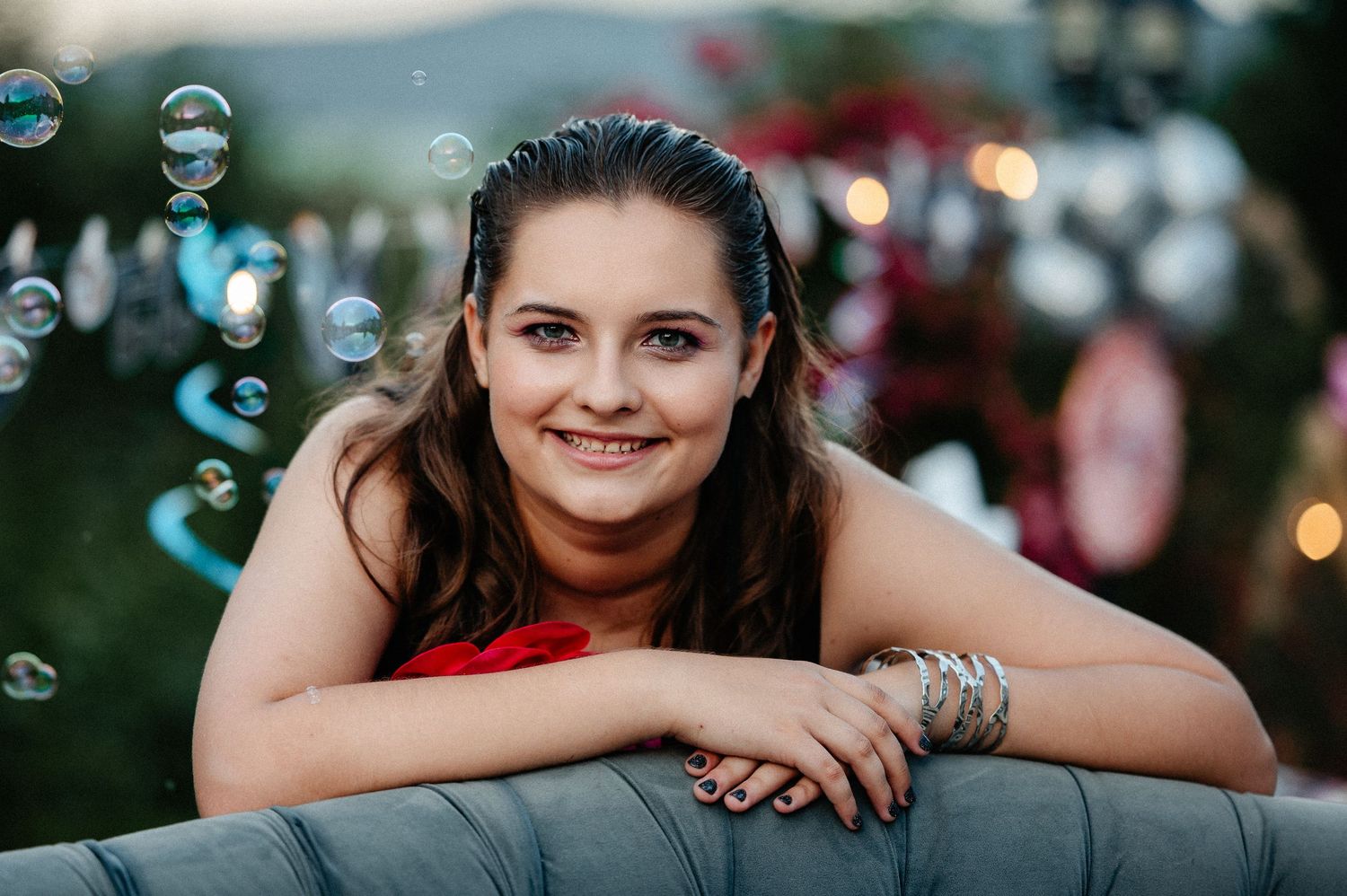 Smiling young woman in red dress leans on gray couch surrounded by floating soap bubbles outdoors.
