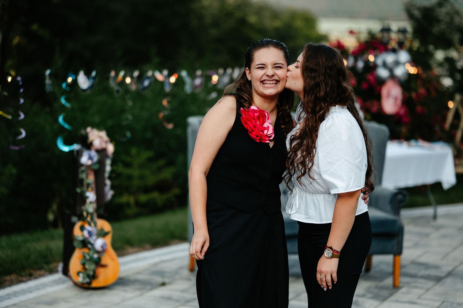 Two young women sharing a tender kiss on the cheek at an outdoor garden celebration.