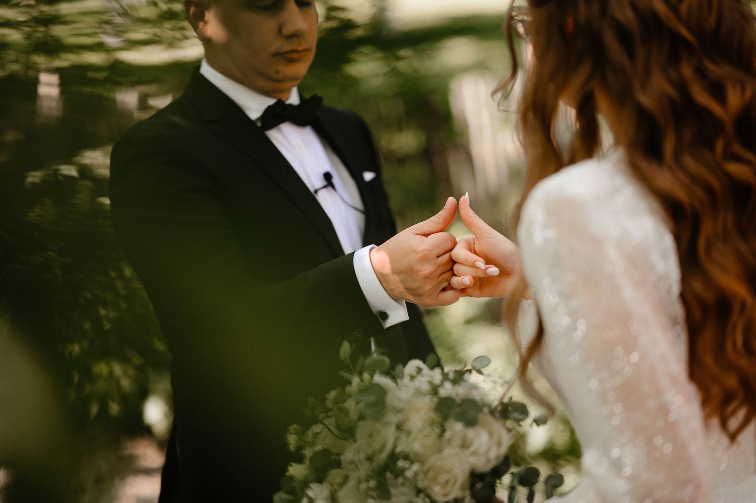 Groom in black tuxedo placing wedding ring on brides finger during outdoor ceremony.
