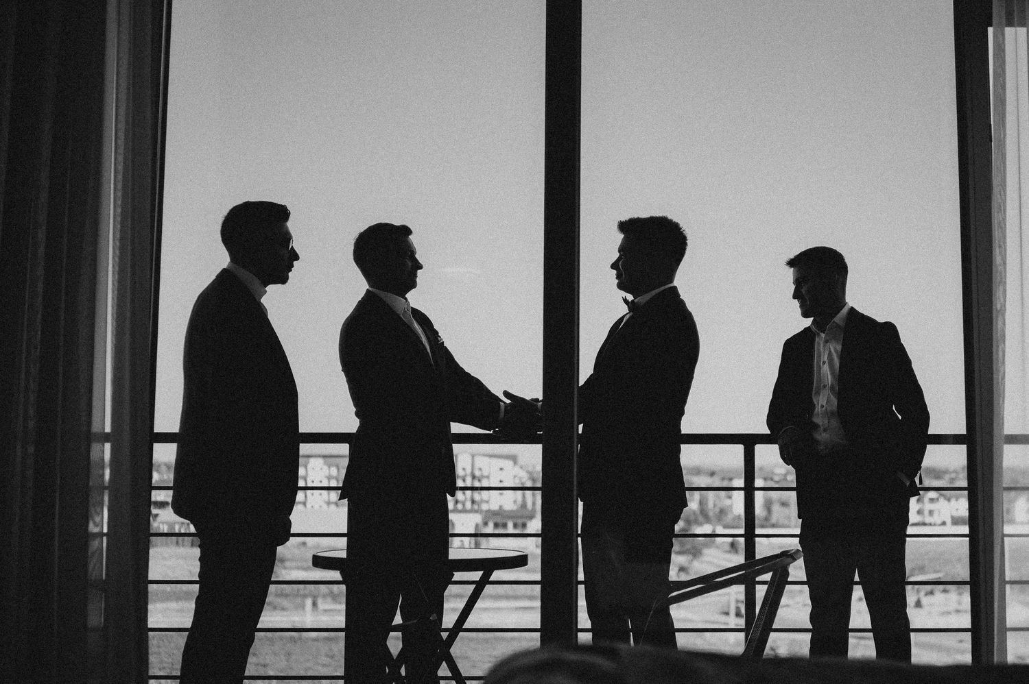 Silhouettes of four men in suits shaking hands by large windows in black and white.