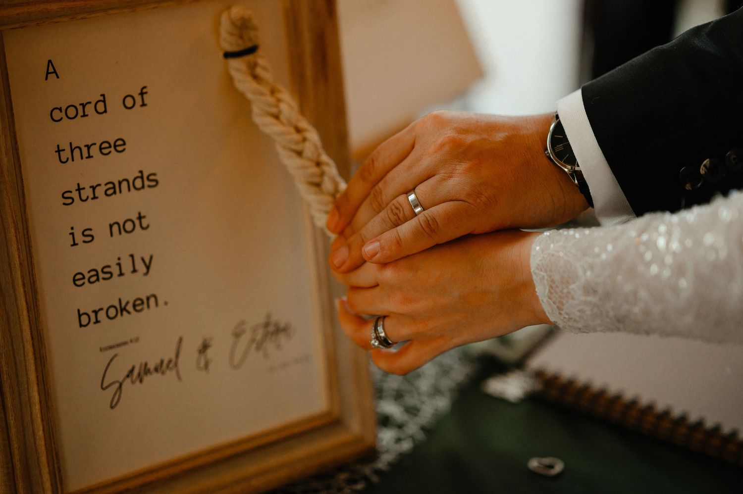 Newlyweds hands with wedding rings rest on a framed sign reading a cord of three strands.