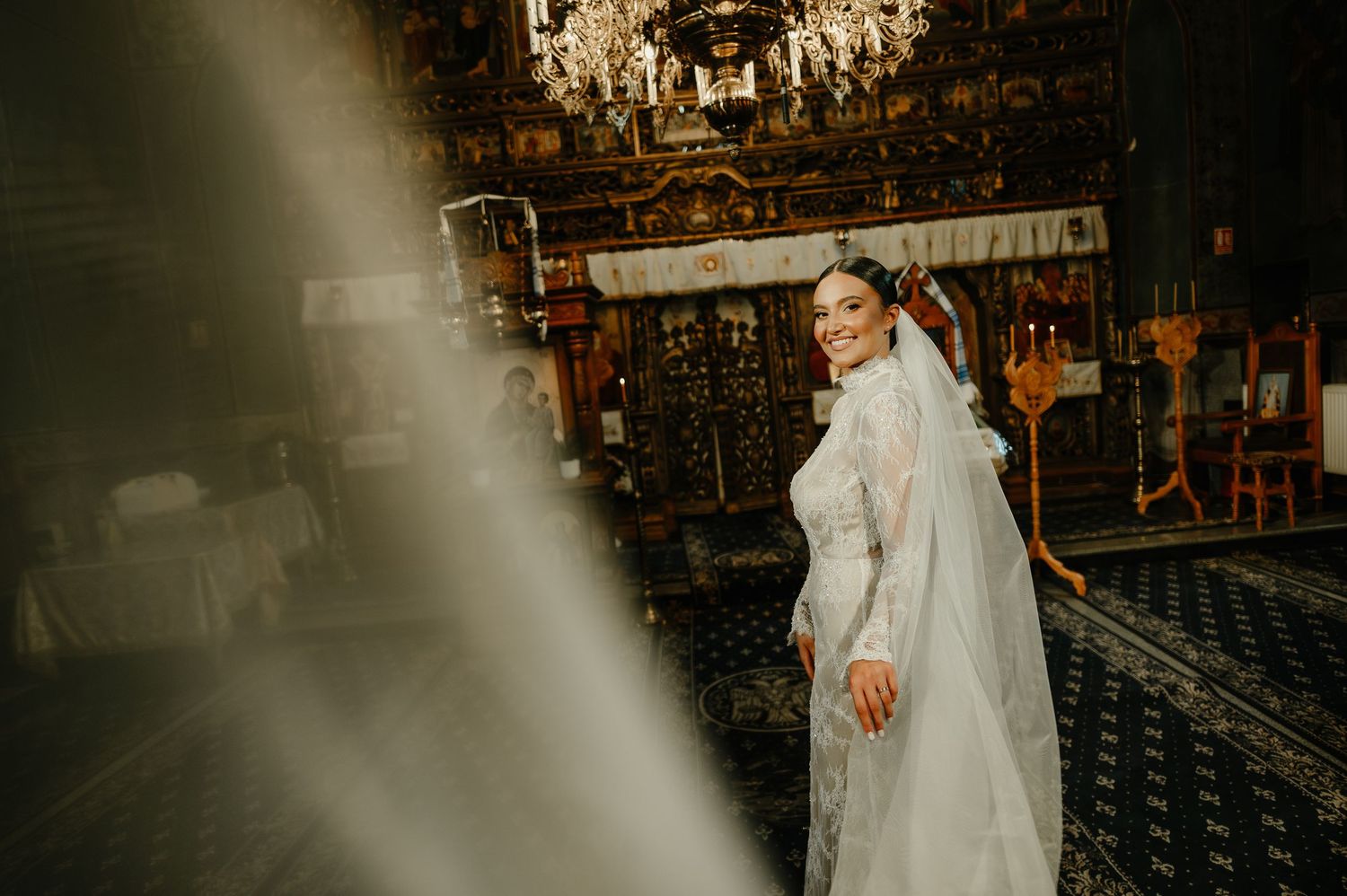 Smiling bride in a long white veil twirling inside an ornate Orthodox church with golden iconostasis.