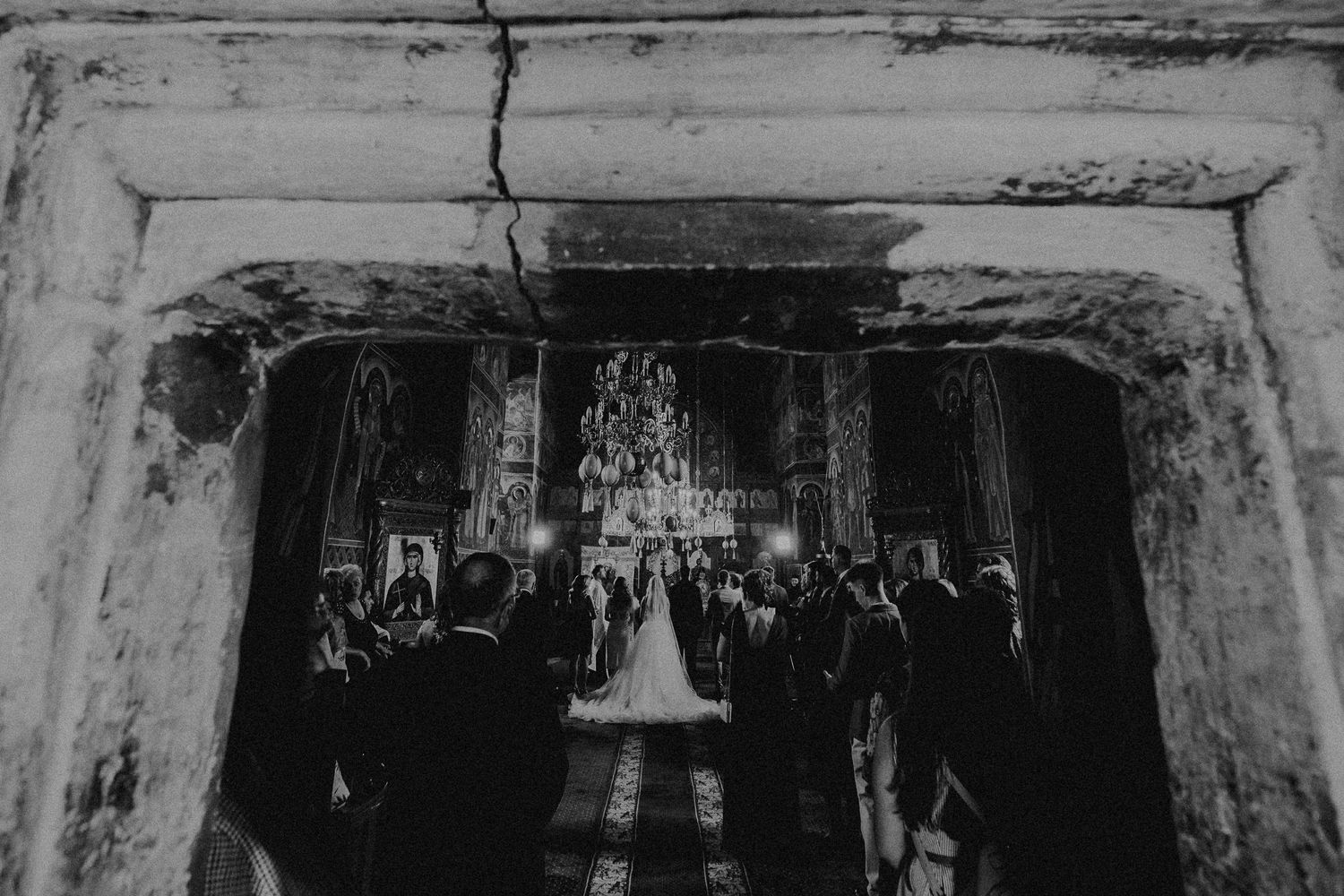 Black and white wide shot of wedding ceremony viewed through stone archway with chandelier and gathered guests.