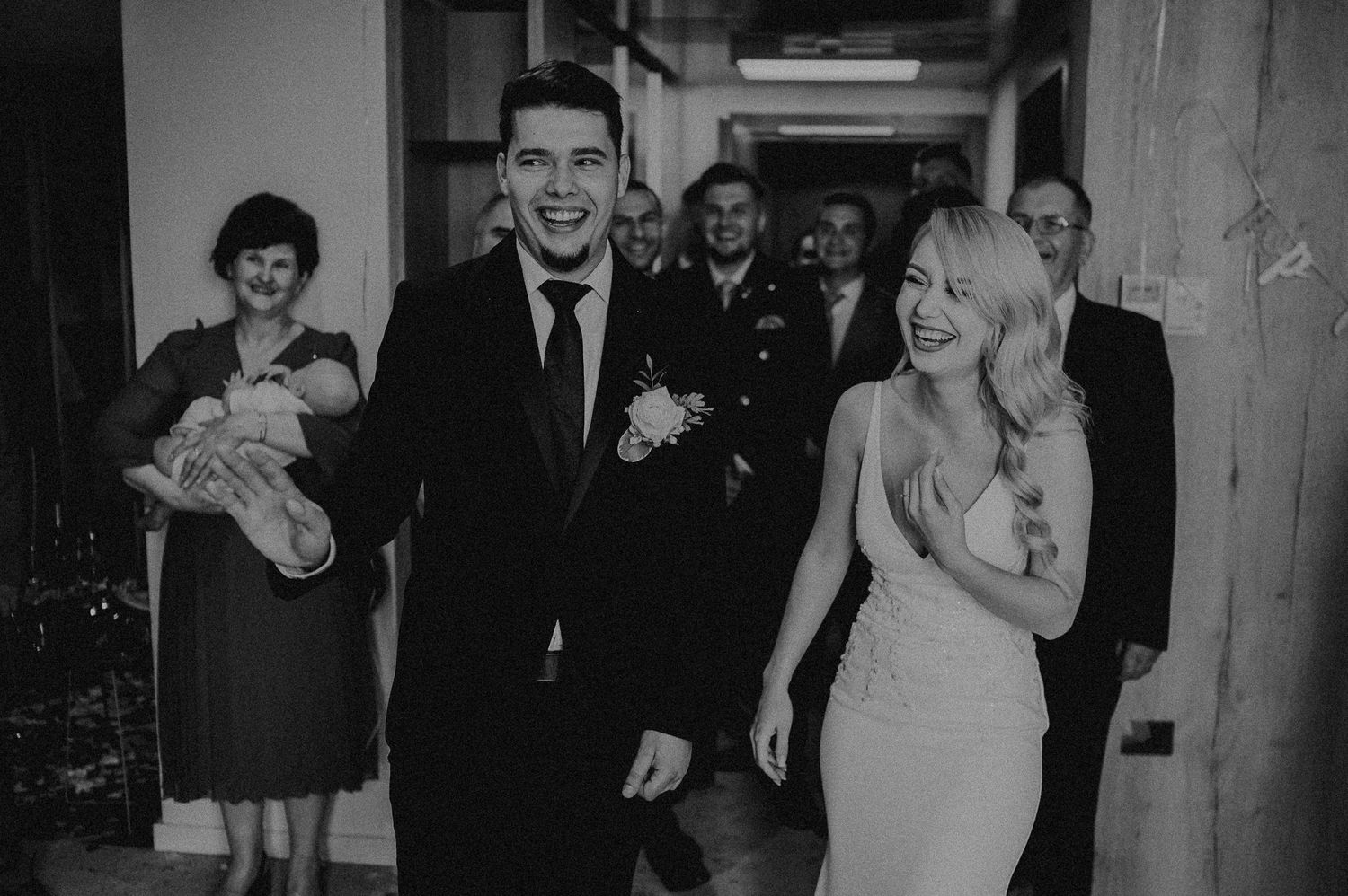 Joyful newlyweds walk down hallway laughing as wedding guests celebrate behind them in black and white photo.