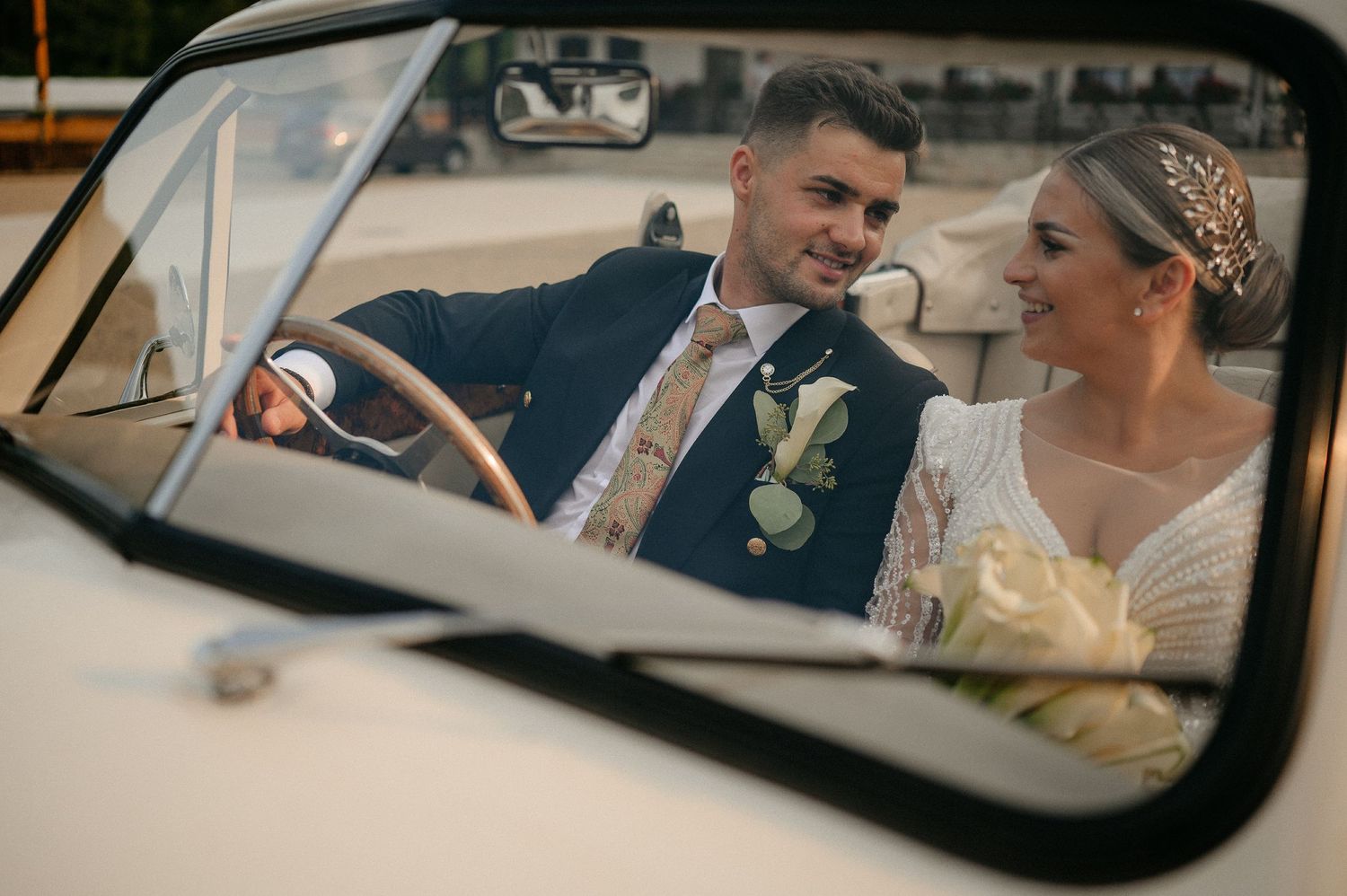 Smiling newlyweds sitting inside a vintage white convertible car on their wedding day.