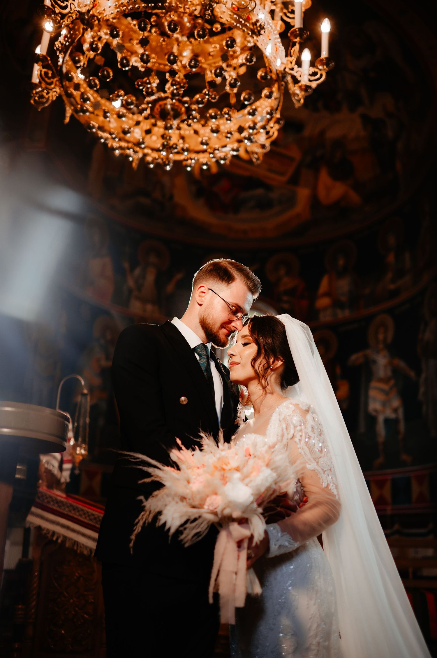 Bride and groom sharing tender moment under ornate chandelier in decorated Orthodox church.