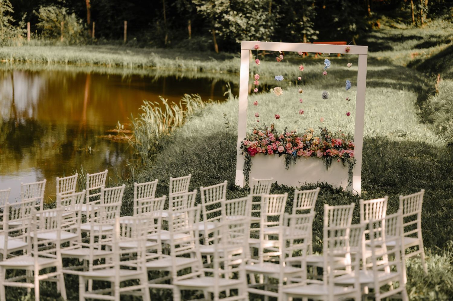 Outdoor wedding ceremony setup with white chairs and floral arch beside a serene lake.