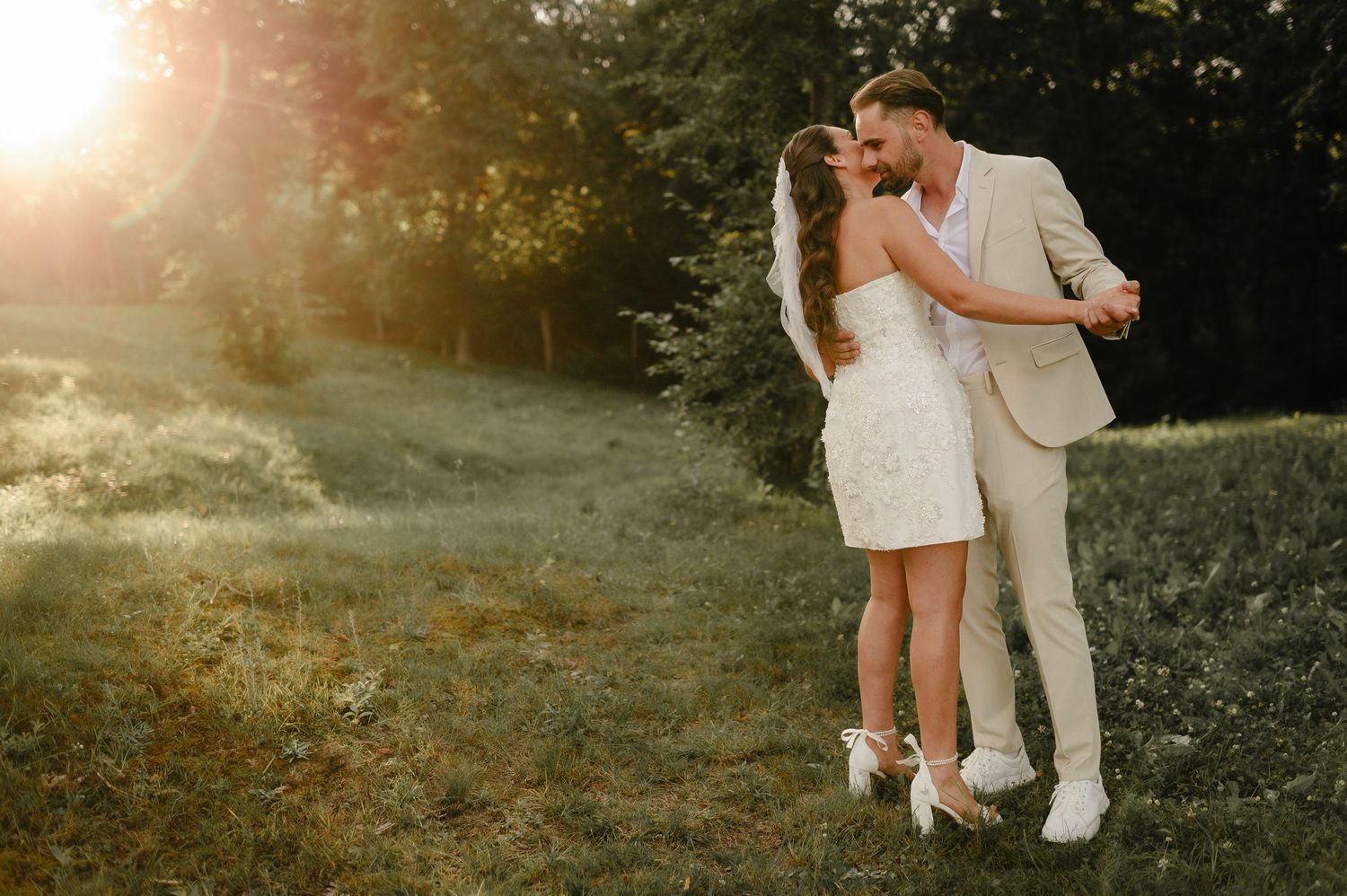 Bride and groom sharing tender kiss in sun-drenched meadow during romantic outdoor summer wedding.
