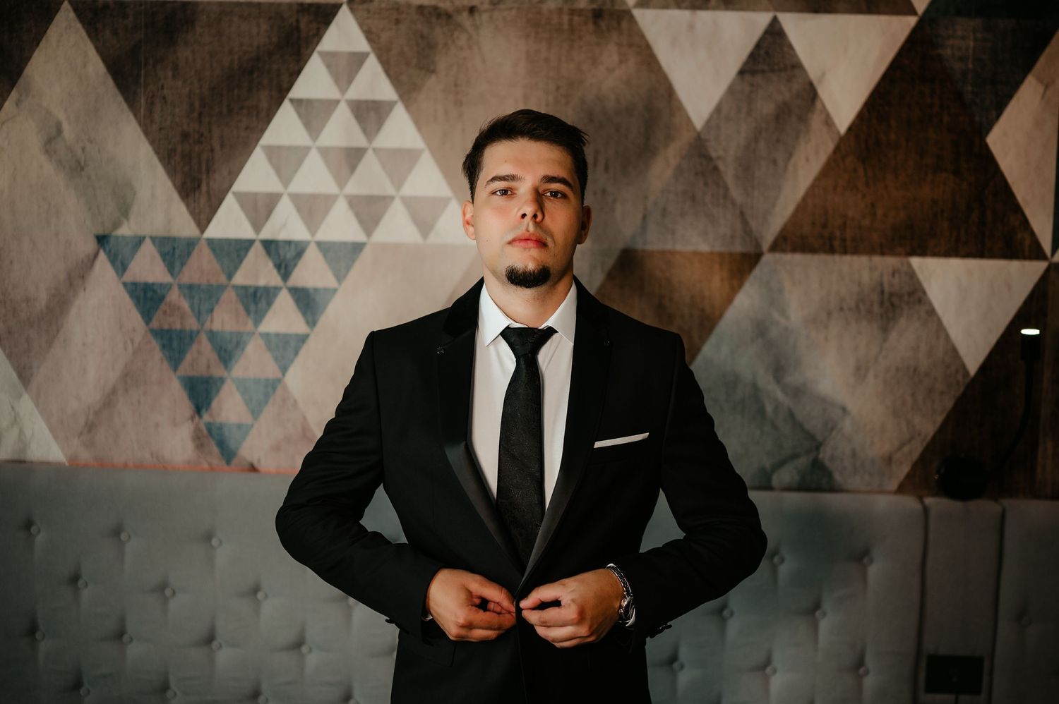 Elegant man in black suit buttoning jacket before a colorful geometric triangle patterned wall.