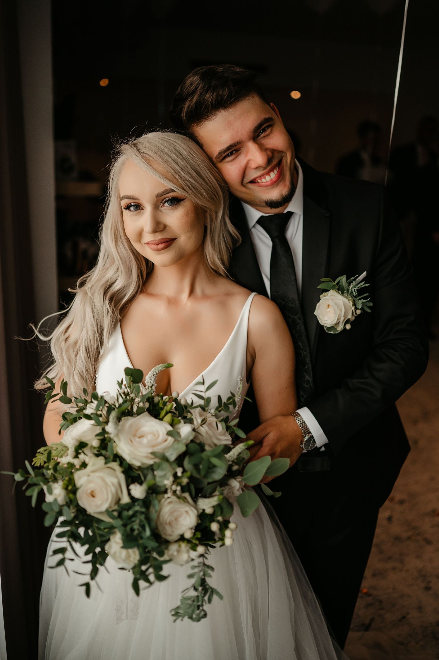 Smiling couple posing together at wedding, bride holding white rose bouquet with lush greenery.