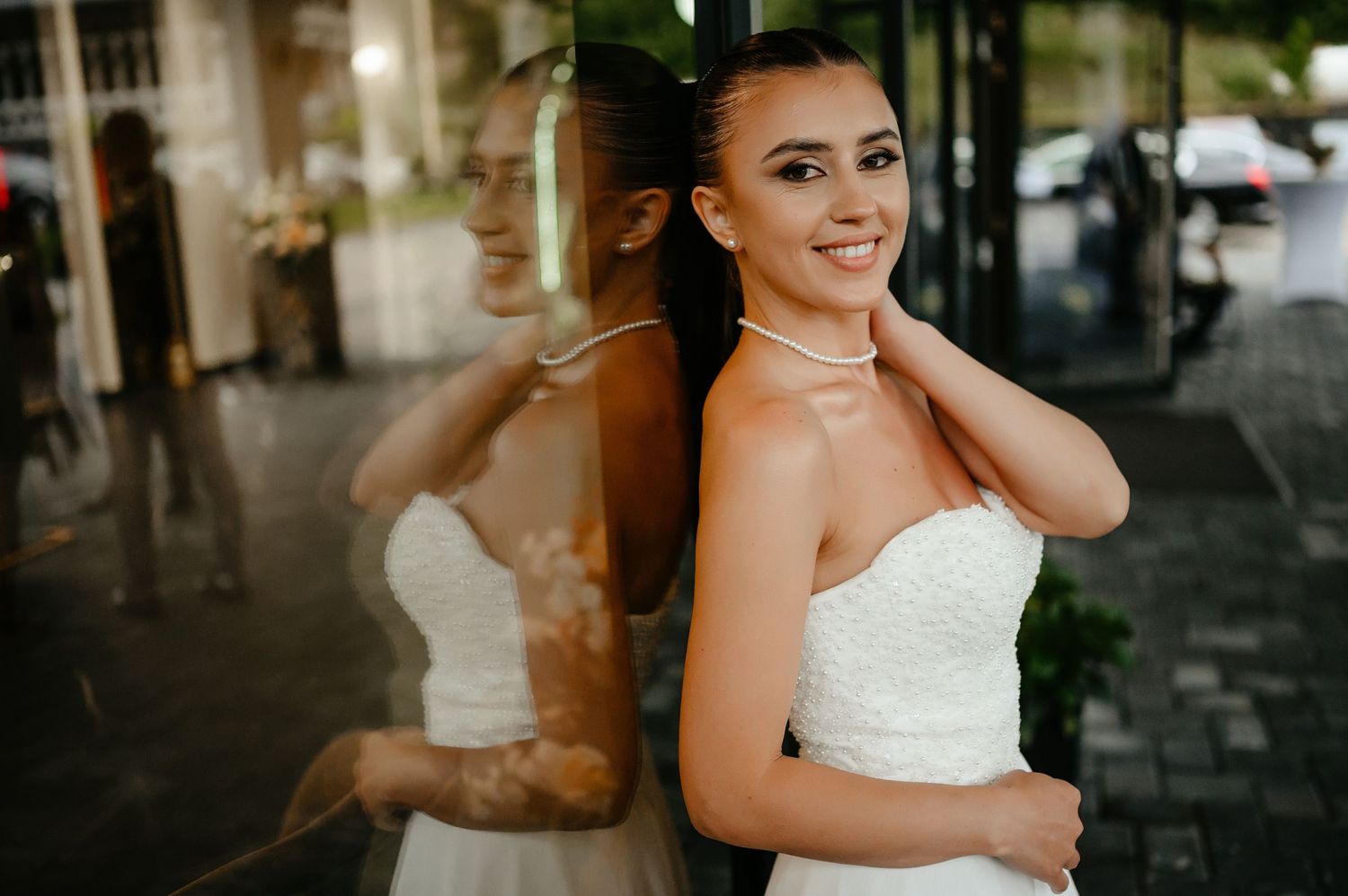 Smiling bride in strapless white dress leaning against glass window with beautiful reflection outdoors.