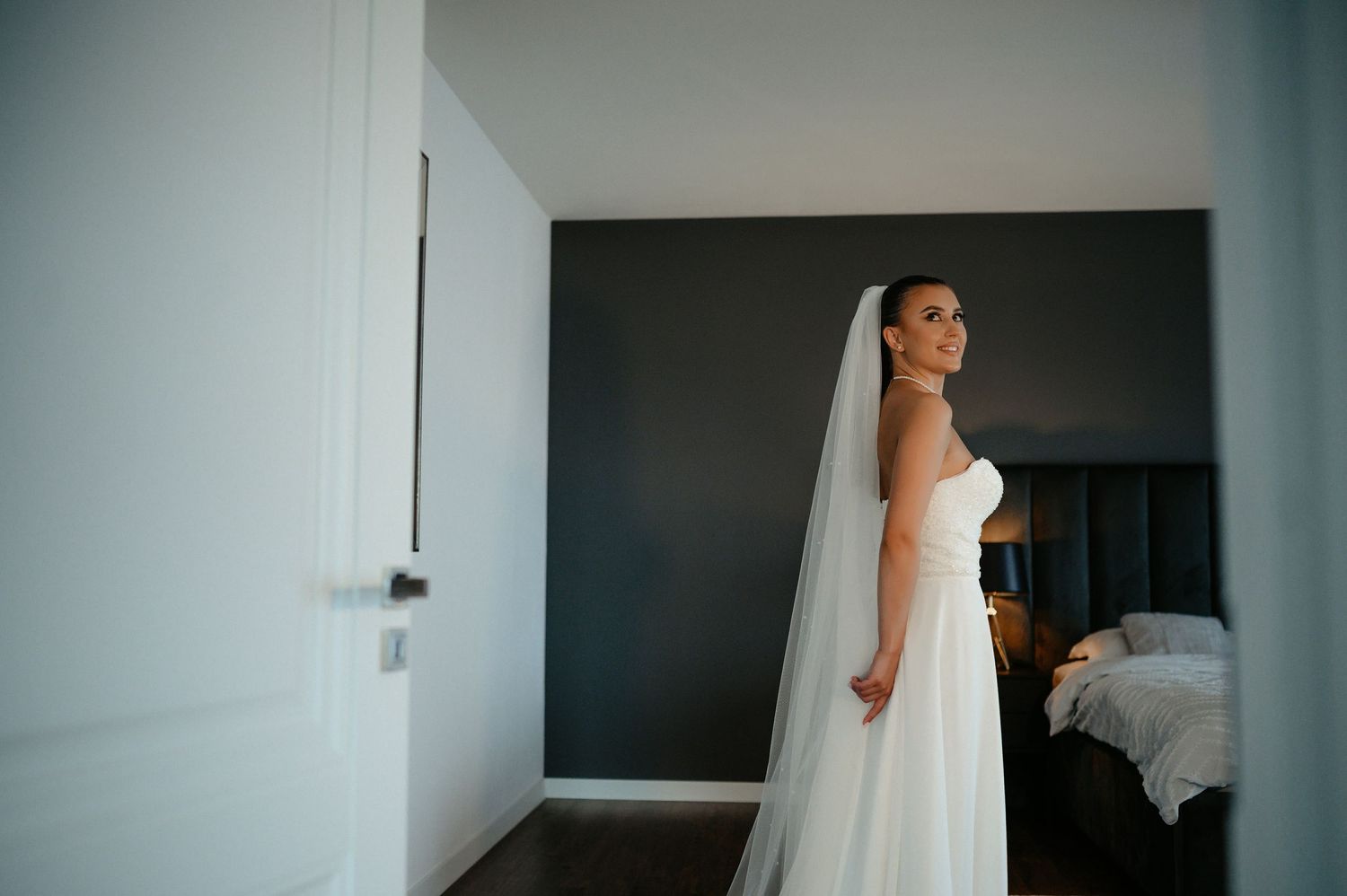 Bride in strapless white gown and veil standing in modern bedroom doorway.