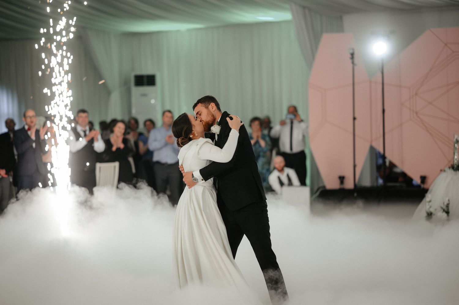 Bride and groom share first kiss during first dance surrounded by dry ice fog and sparklers.