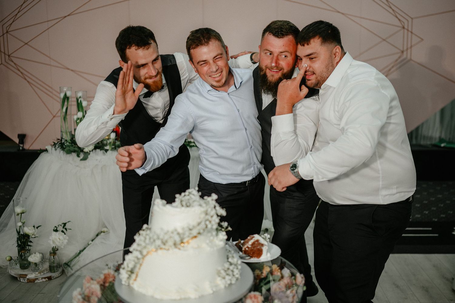 Groom poses with groomsmen cutting white wedding cake together in fun celebratory wedding reception moment.