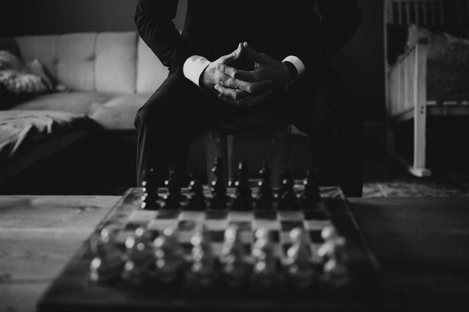 Black and white close-up of a groom in a tuxedo contemplating a chess board on a wooden table.