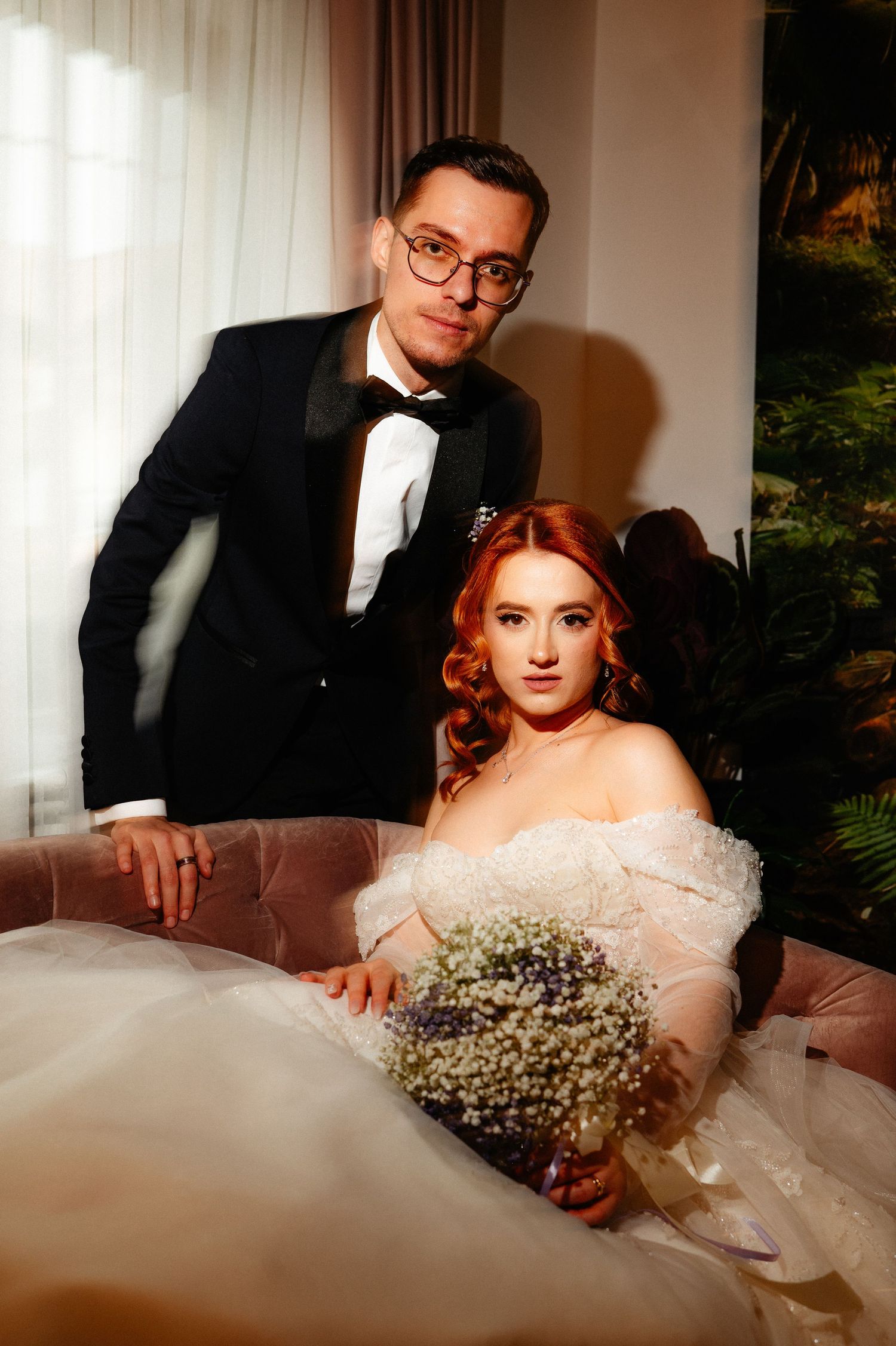 Bride in off-shoulder gown holds baby's breath bouquet while groom in tuxedo leans beside her on sofa.