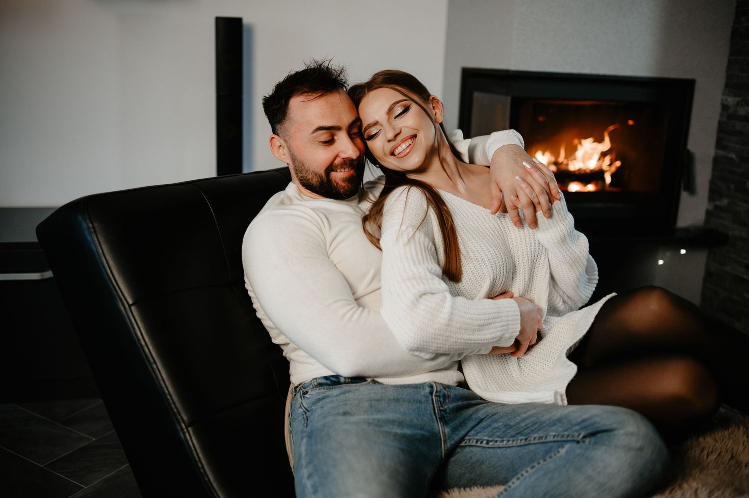 Happy couple in white sweaters sit together embracing warmly in front of a cozy fireplace.