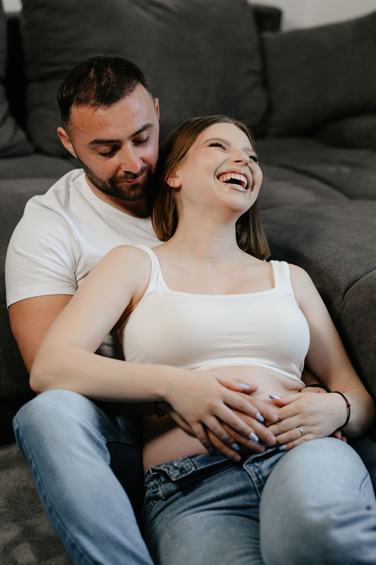 Happy pregnant woman laughing with partner sitting on floor against sofa, both holding baby bump together.