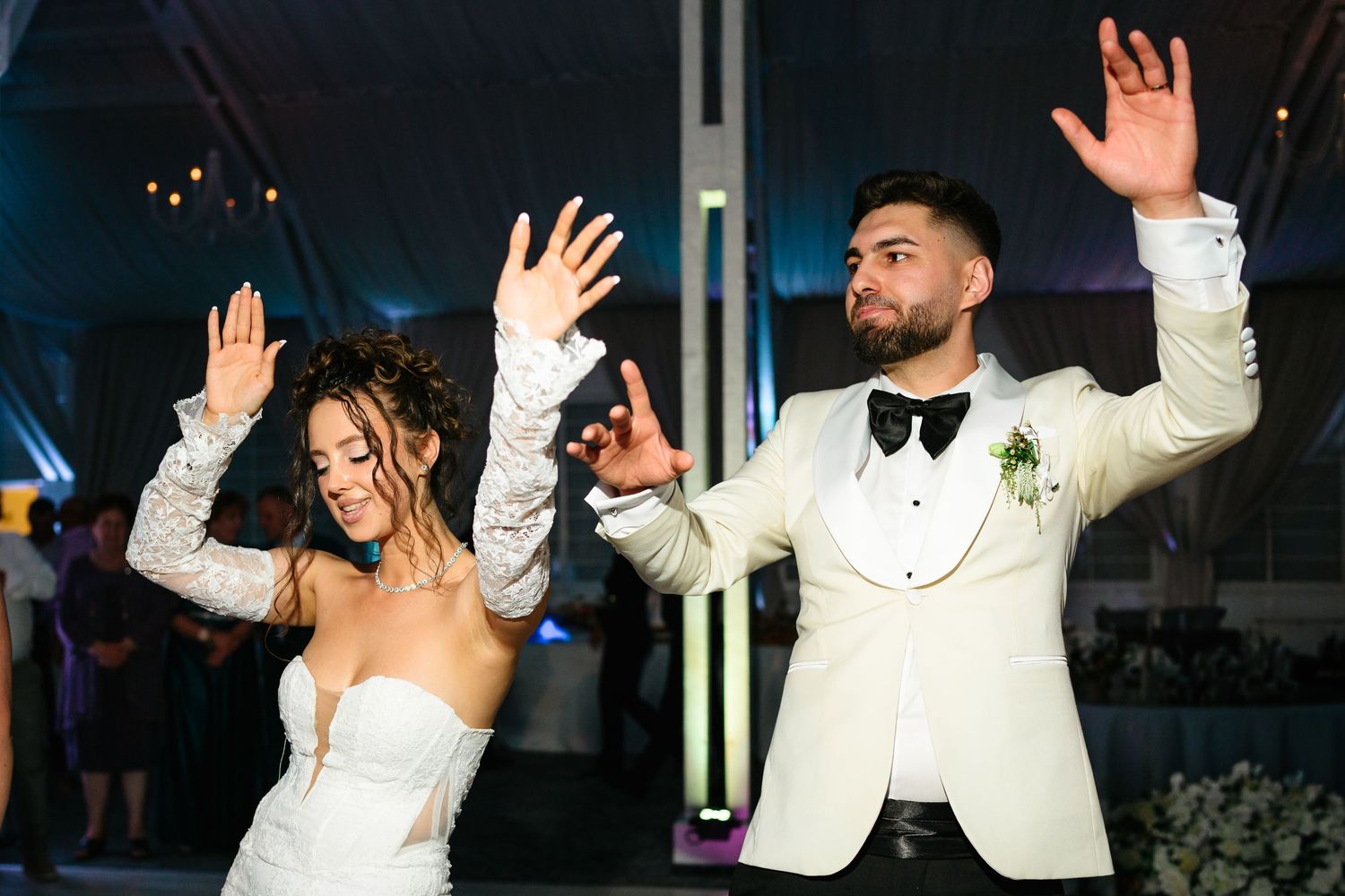 Bride and groom dancing joyfully at wedding reception, both raising hands, groom in white tuxedo with boutonniere.