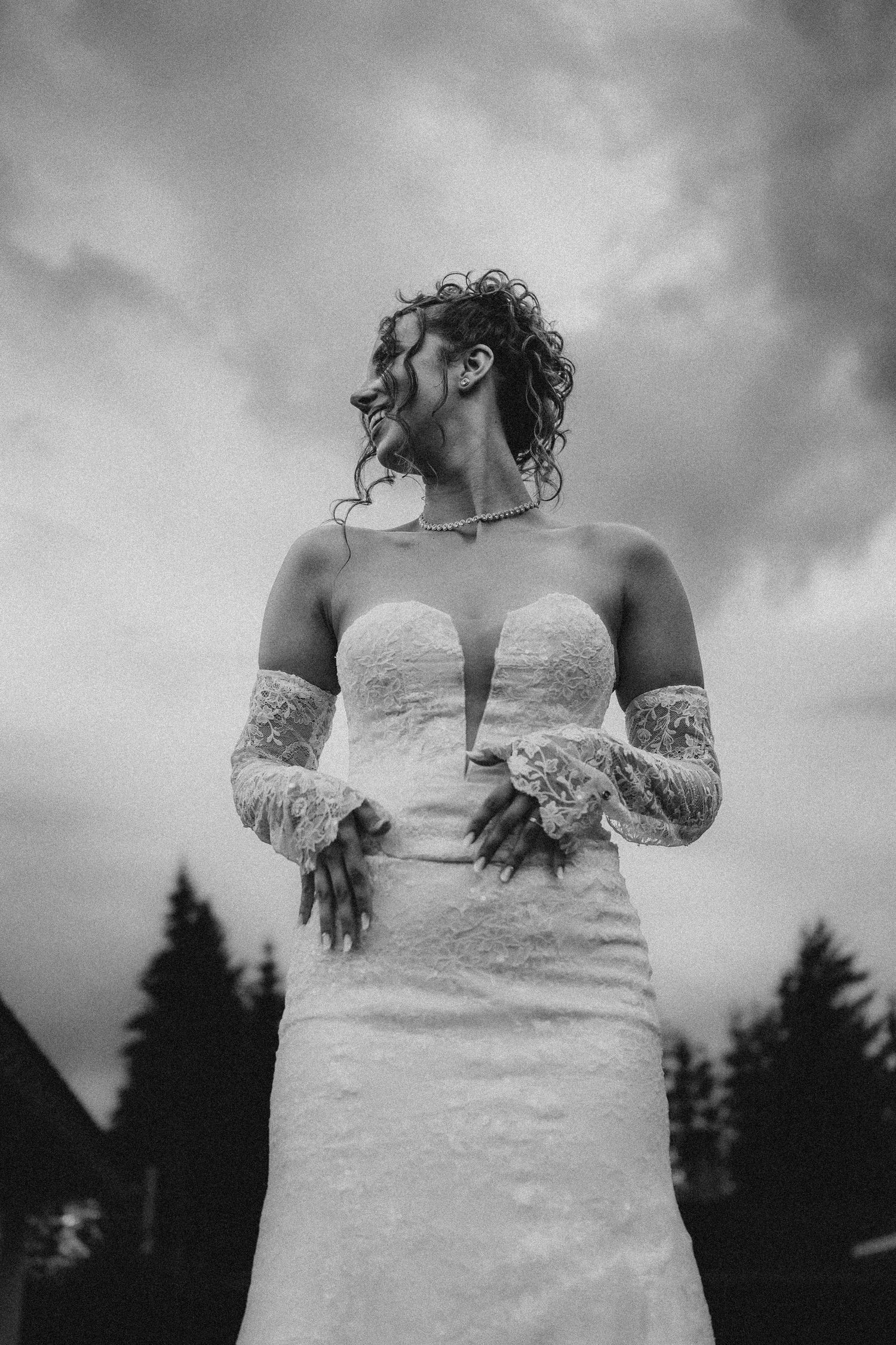 Black and white photo of a bride in a strapless lace gown with lace gloves, smiling outdoors under a cloudy sky.