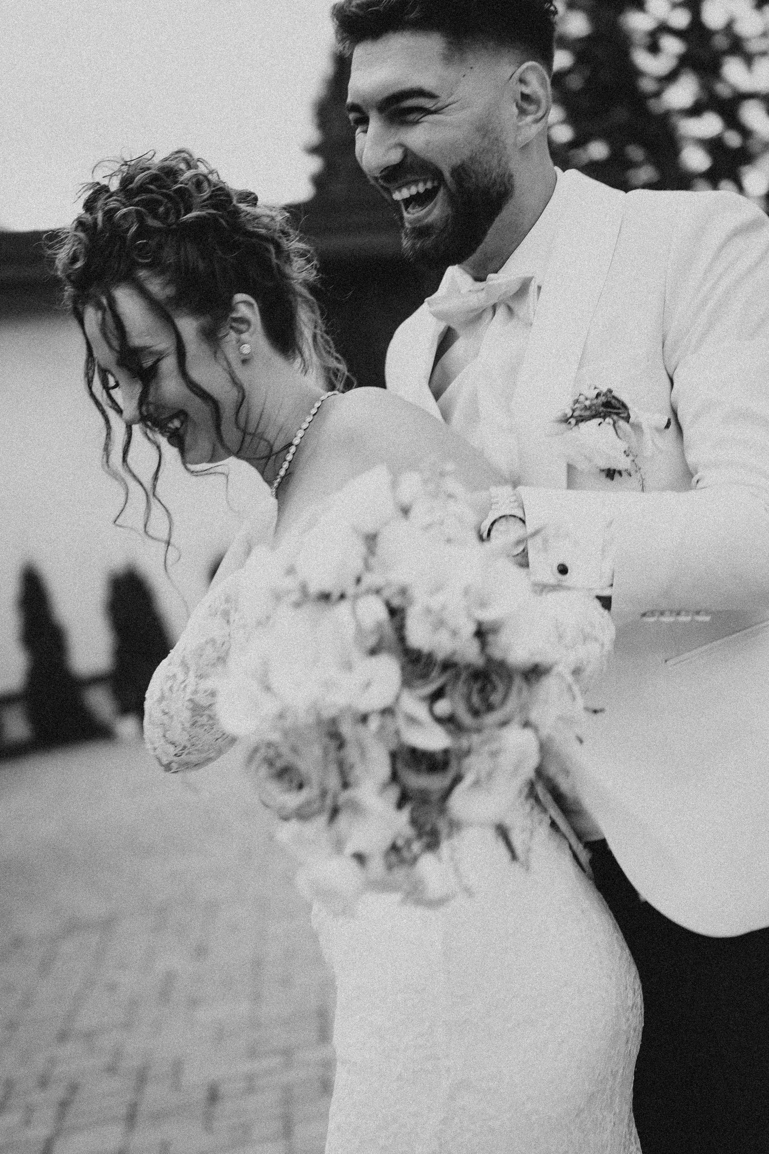 Black and white photo of a laughing bride and groom embracing, bride holding a floral bouquet on their wedding day.