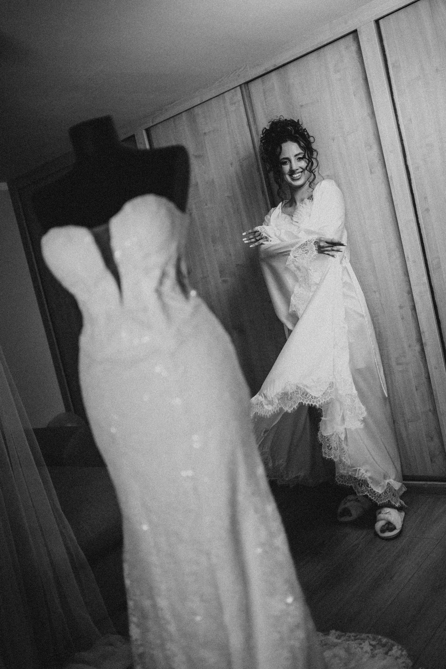 Black and white photo of a smiling bride in a lace wedding dress holding her gown while standing in a room.