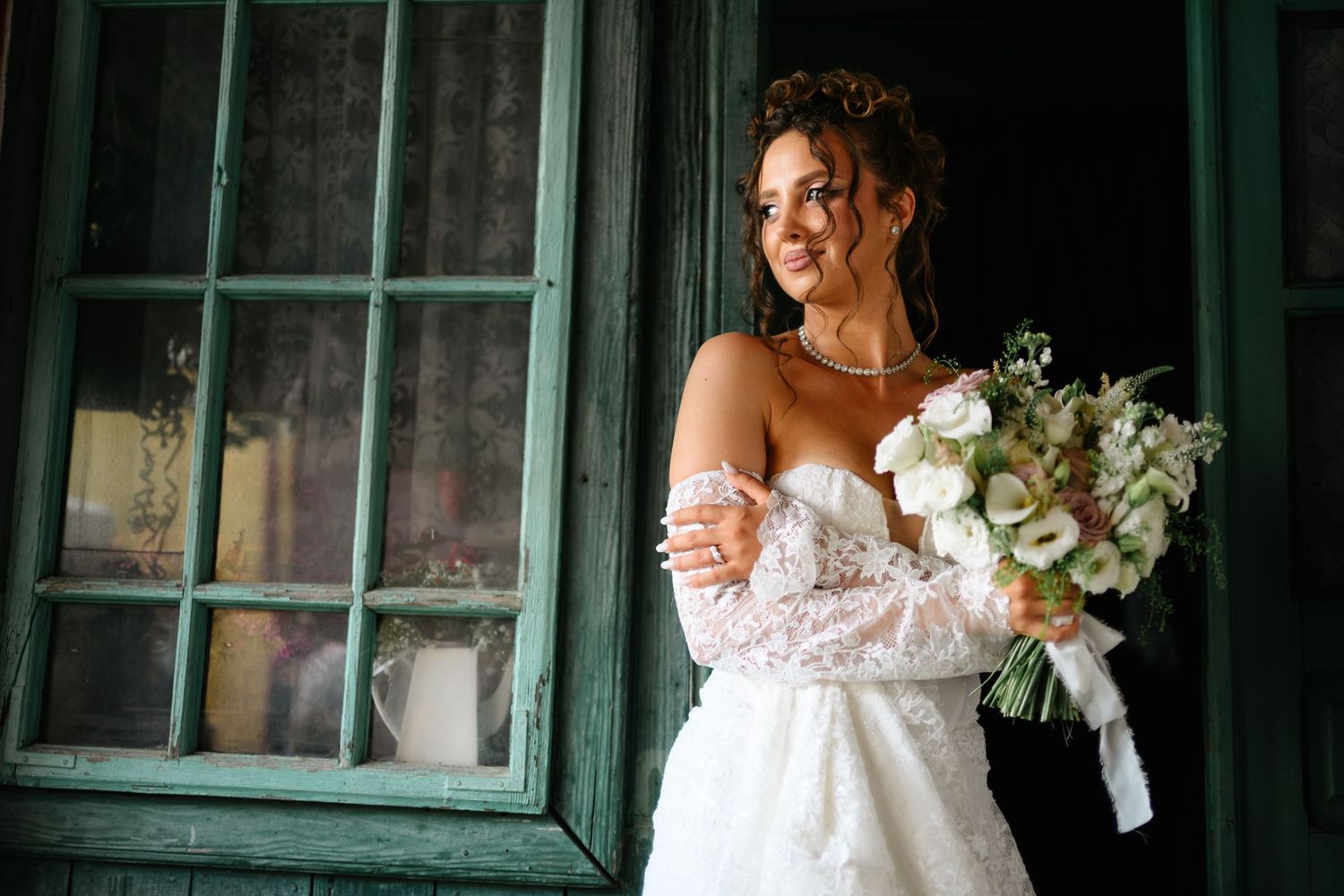 Bride in white lace gown holding elegant bouquet of white flowers, standing by rustic green door.