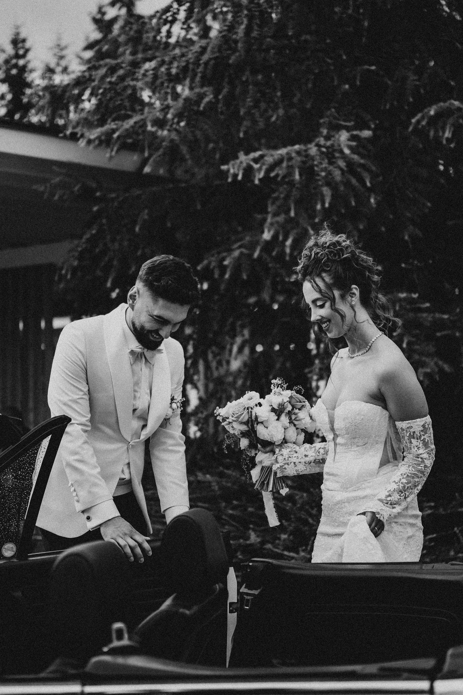 Black and white photo of a bride holding bouquet and groom in white suit beside a vintage car outdoors.