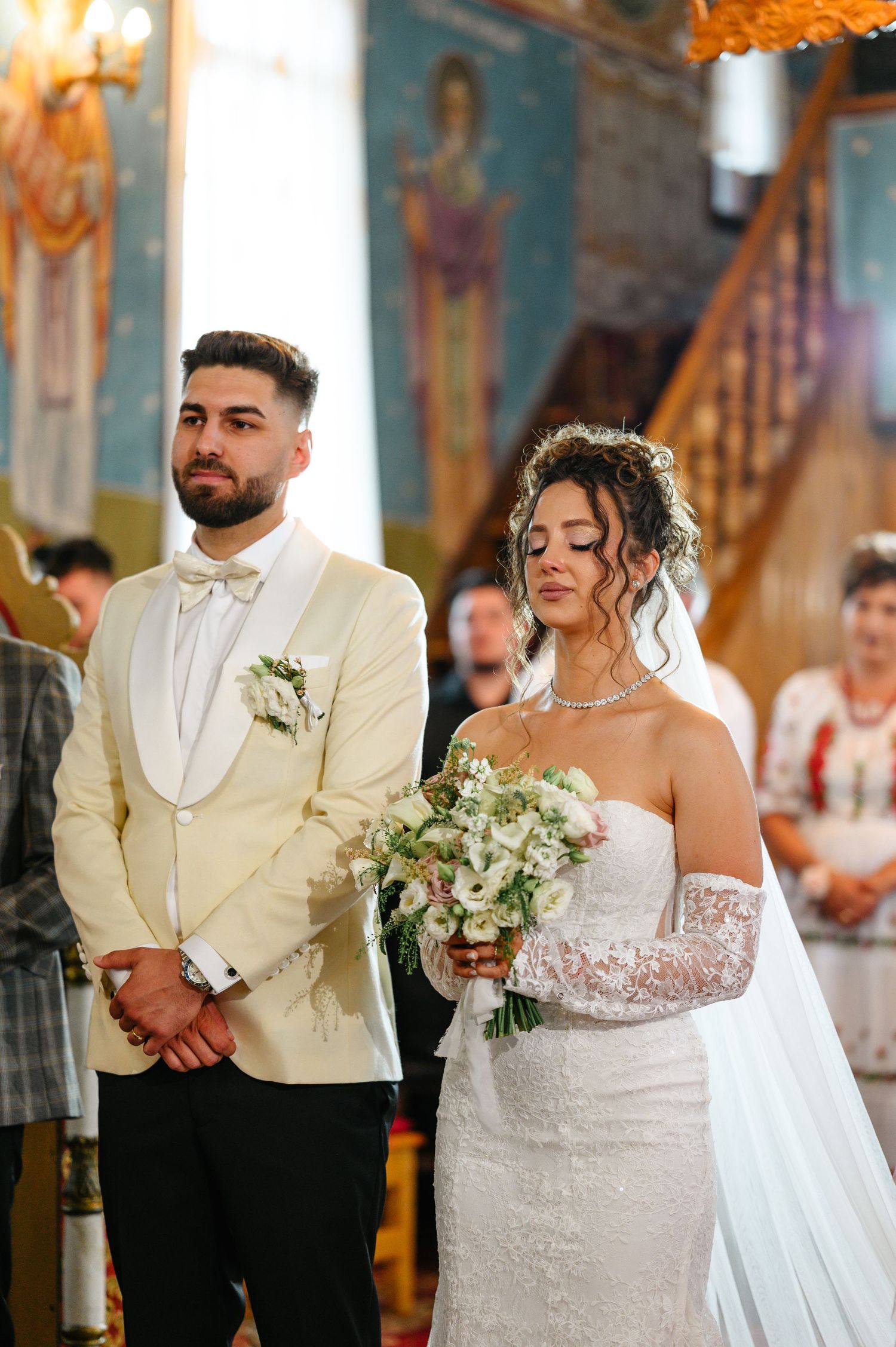 Bride and groom stand at church altar, she holds white bouquet in lace gown, he wears cream tuxedo with bow tie.