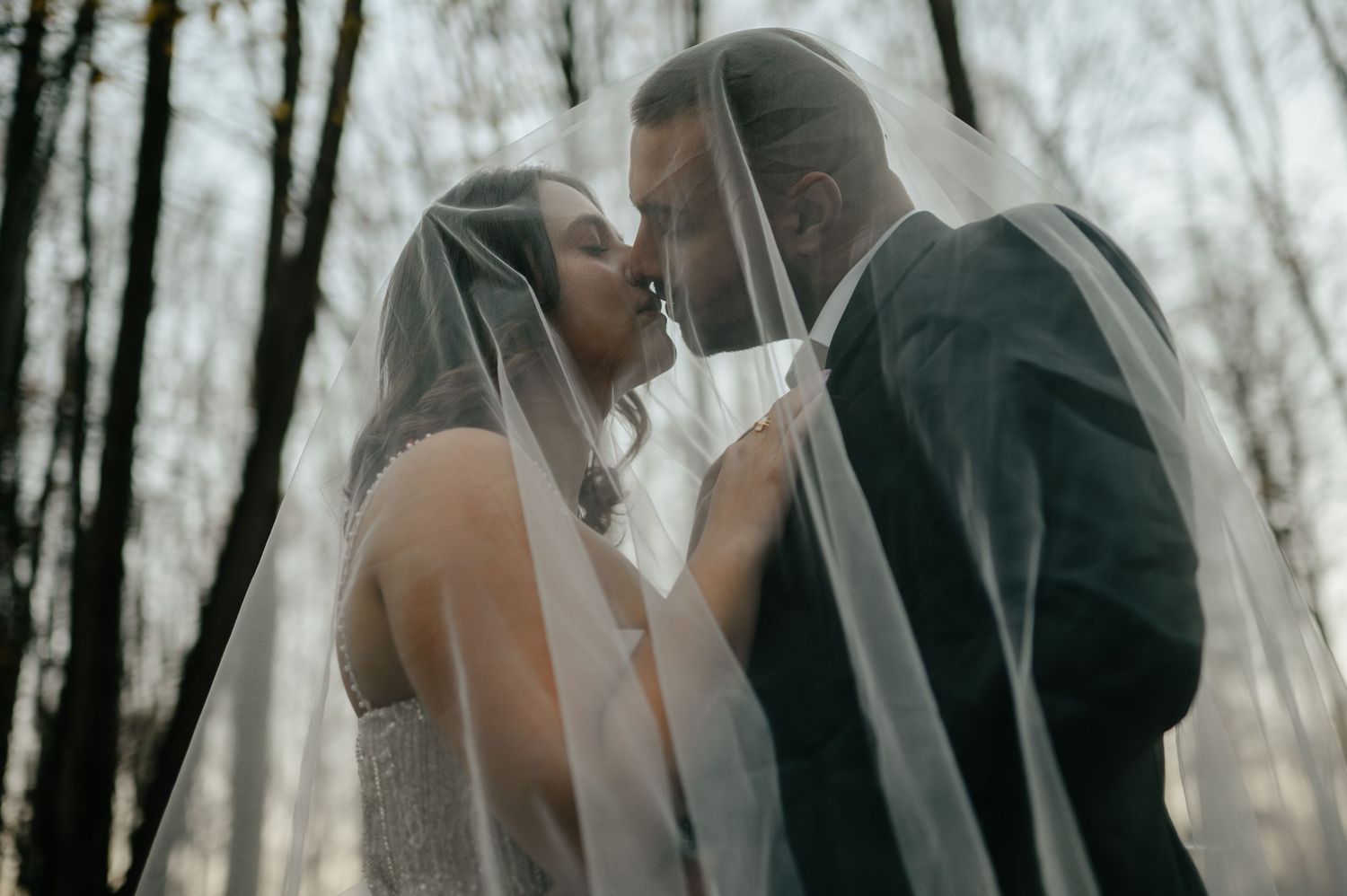 Bride and groom share a romantic kiss under a flowing wedding veil in a forest setting.