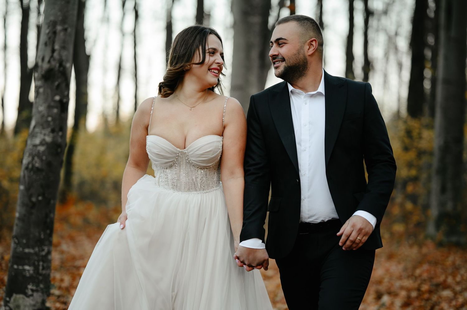 A bride and groom walk hand in hand through an autumn forest, smiling at each other in wedding attire.