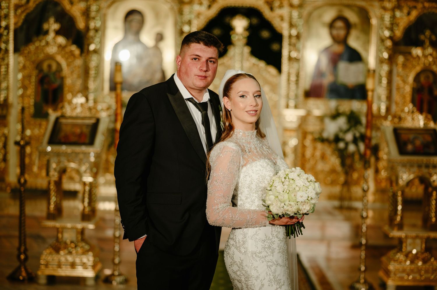 Bride and groom posing in an ornate Orthodox church with golden icons, bride holding white bouquet.