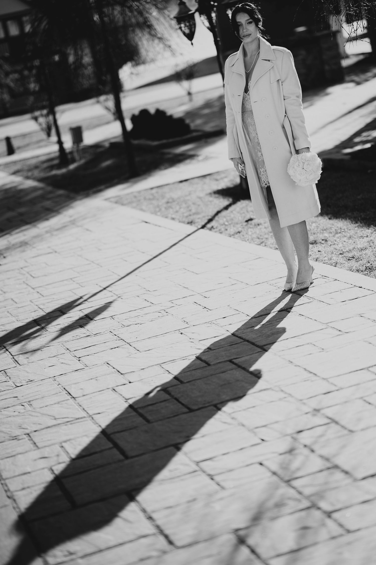 Black and white photo of bride in long white trench coat standing on sunlit stone pathway casting long shadow.