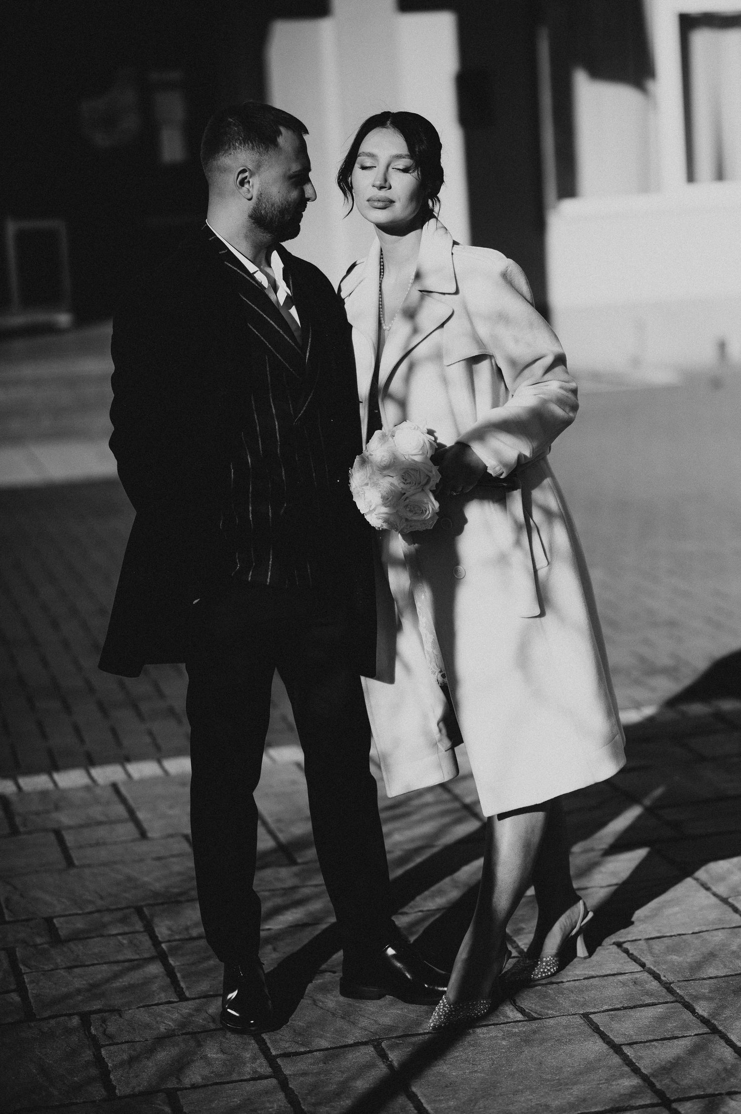 Elegant couple in black and white photo, man in pinstripe suit and woman in long coat on cobblestone street.