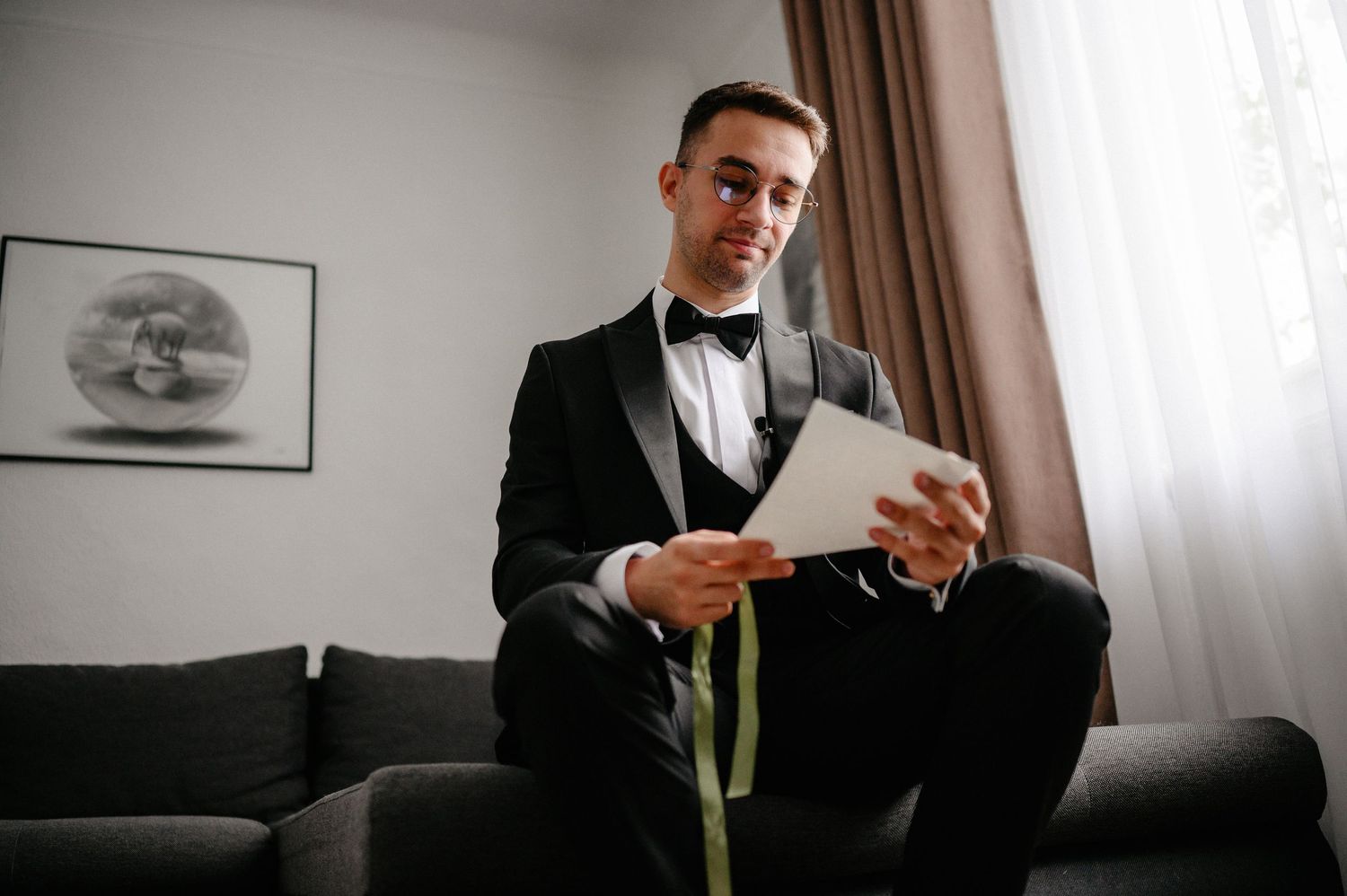 Young man in tuxedo sitting on sofa reading a card, dressed for formal event with bow tie and boutonniere.