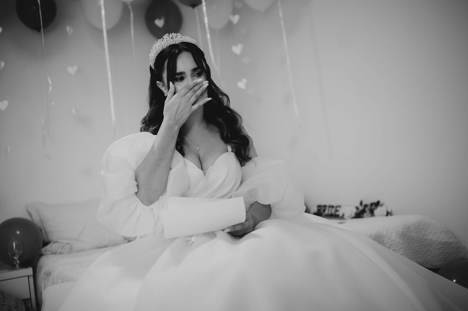 Emotional bride in white gown and tiara covering her face while sitting on bed with heart decorations.