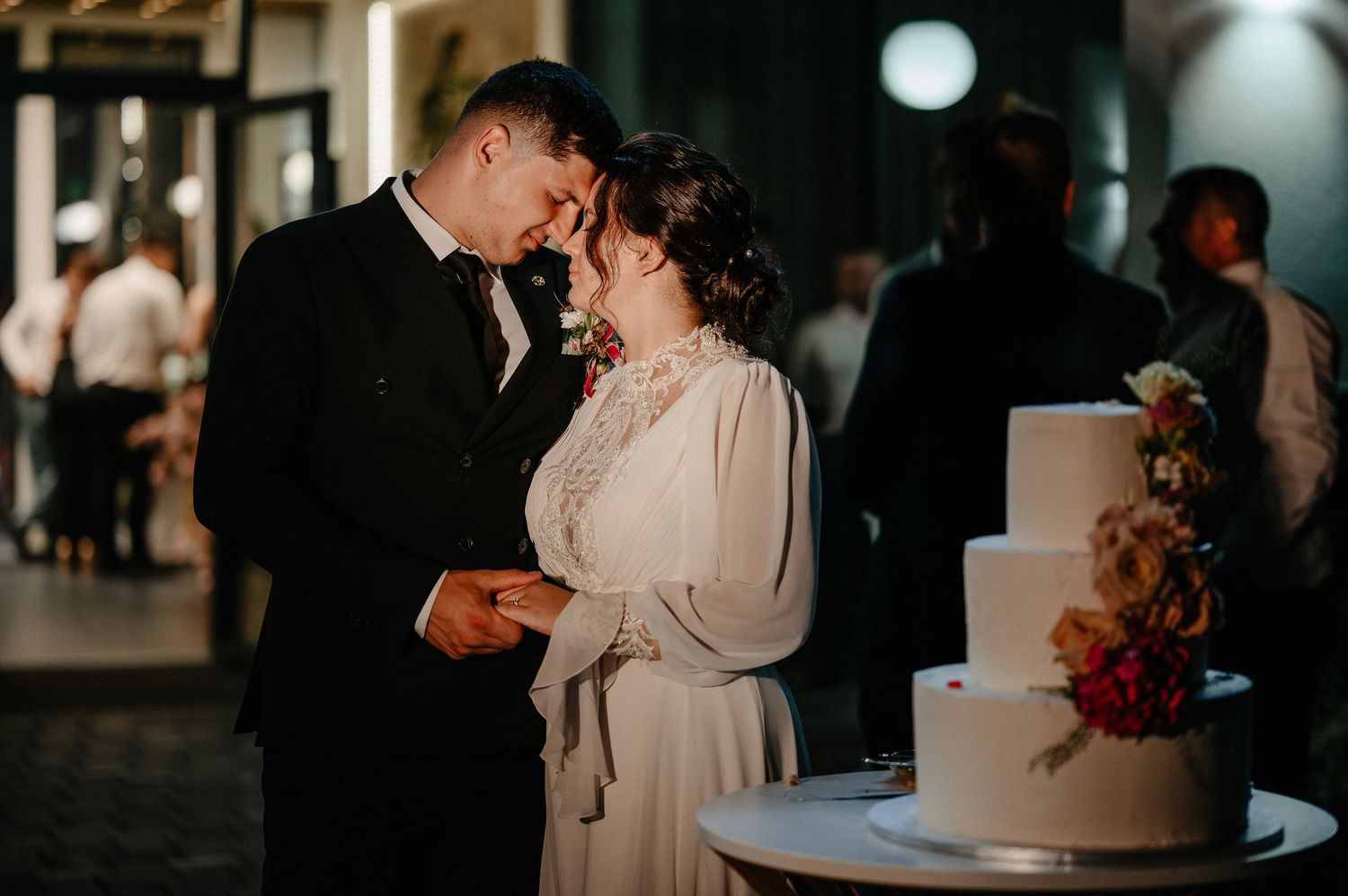 Bride and groom share romantic moment beside floral wedding cake at reception.