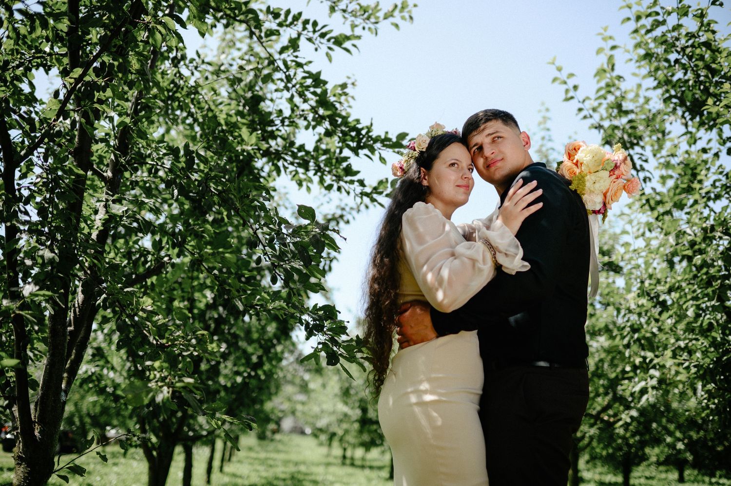 Couple embracing in an orchard, bride wearing floral crown holding bouquet, groom in black suit, lush green trees.