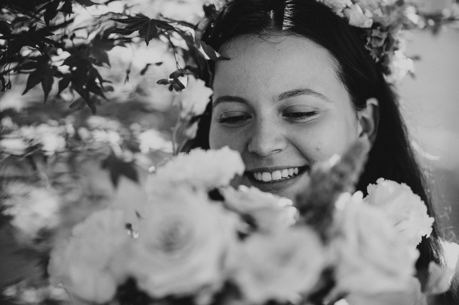 Black and white portrait of a smiling woman surrounded by blooming flowers wearing a floral crown.