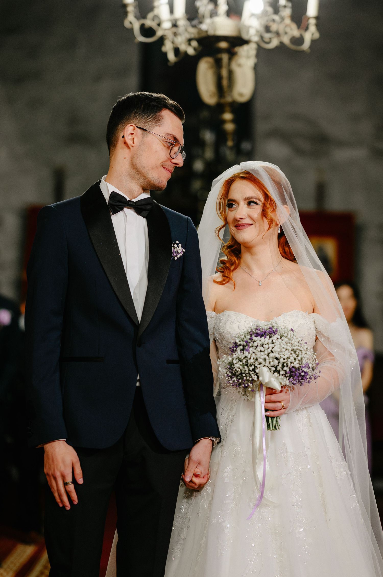 Elegant bride and groom holding hands at wedding ceremony, bride in strapless gown with veil holding purple bouquet.