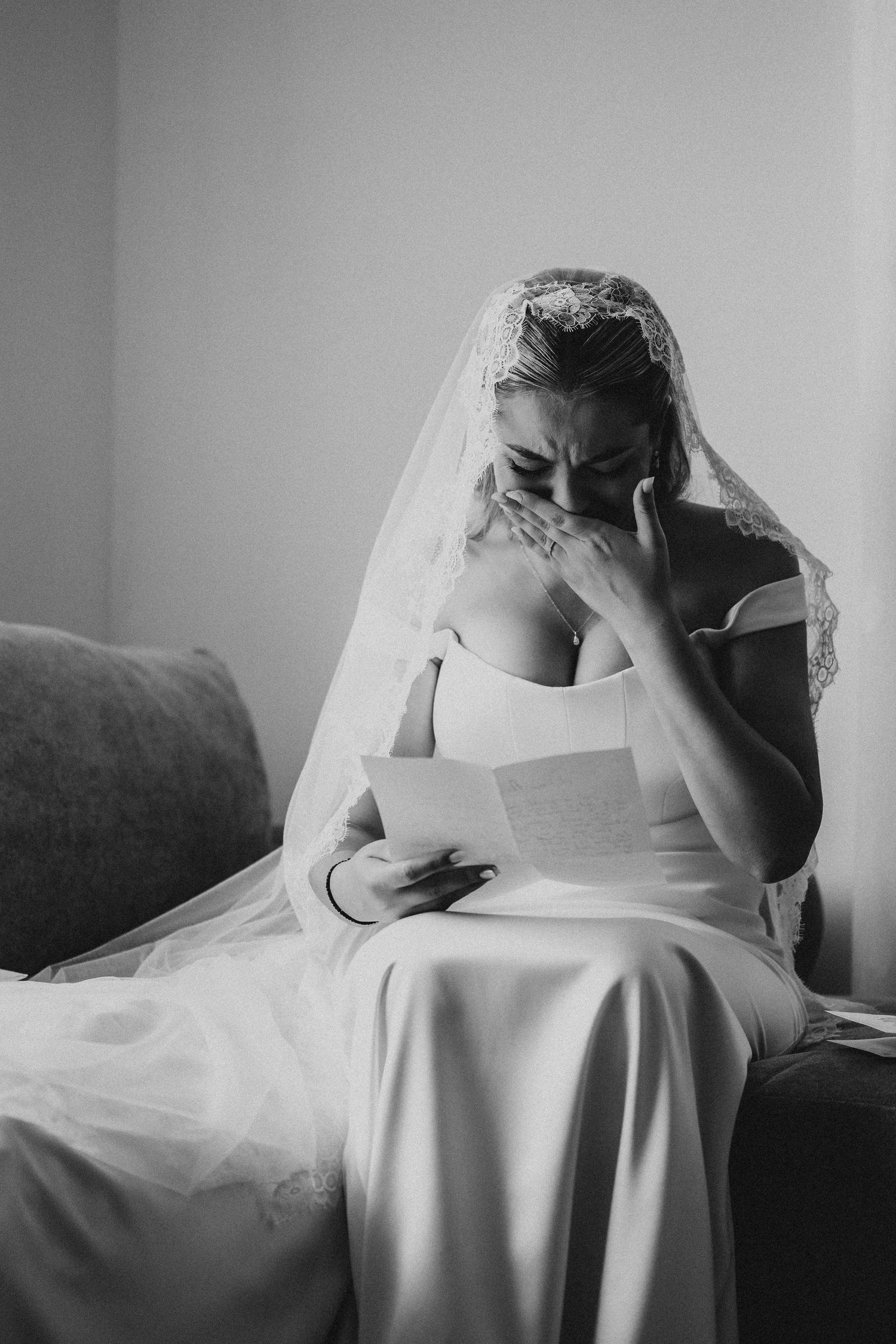Black and white photo of an emotional bride in wedding dress and veil reading a letter while wiping tears on her wedding day.