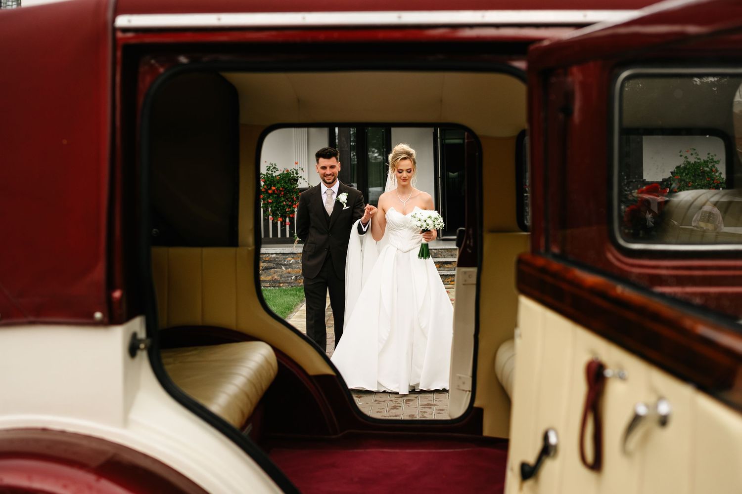 Bride and groom reflected in vintage wedding car window, bride in white gown holding bouquet.