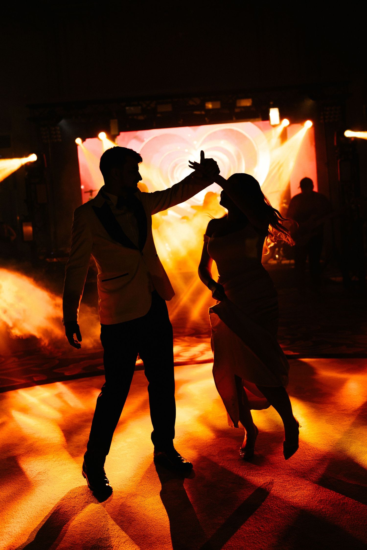 Silhouettes of a couple dancing romantically on a dance floor with dramatic orange and golden stage lights glowing behind them.