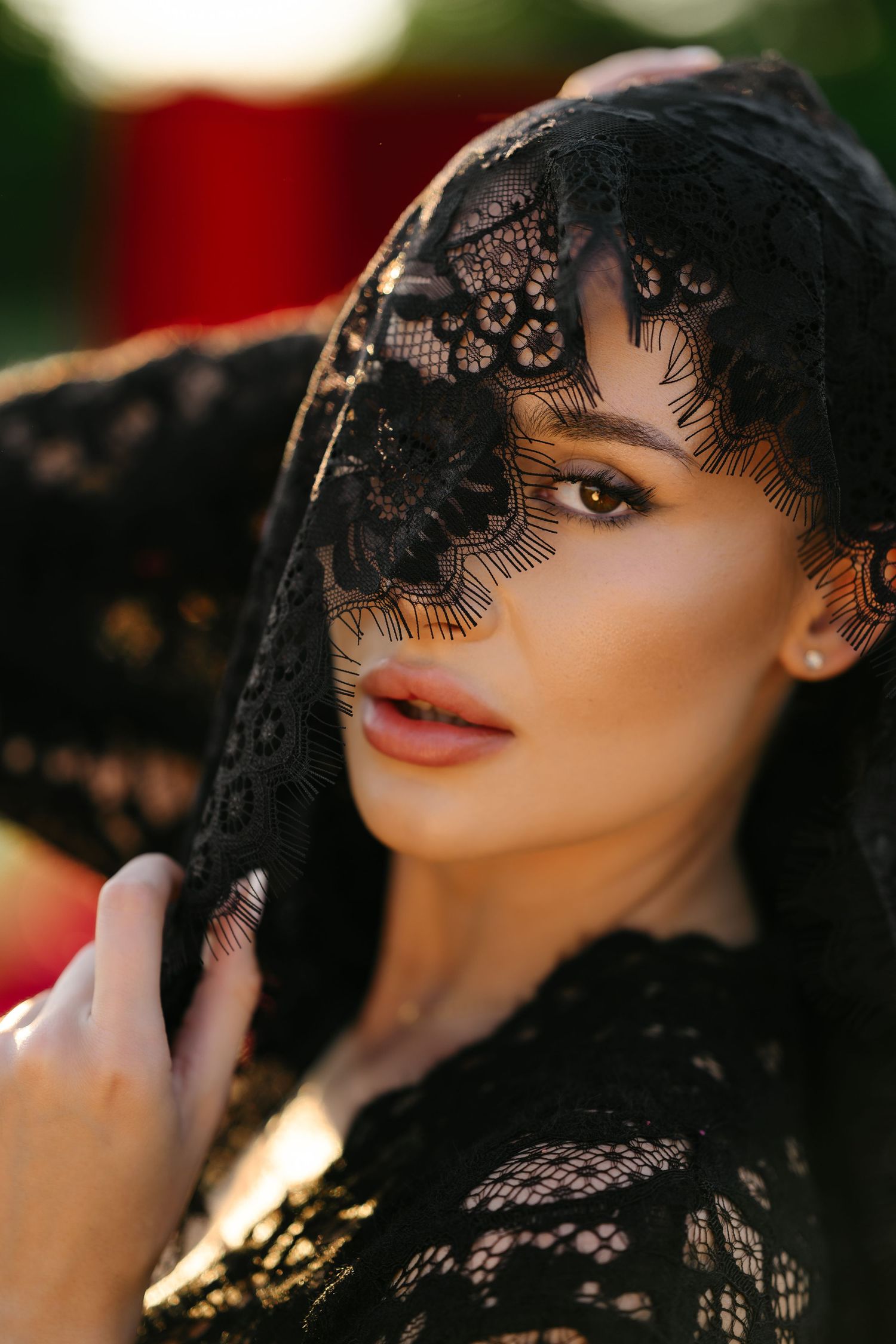 A woman in black lace veil covering her face, gazing seductively at the camera in warm outdoor lighting.