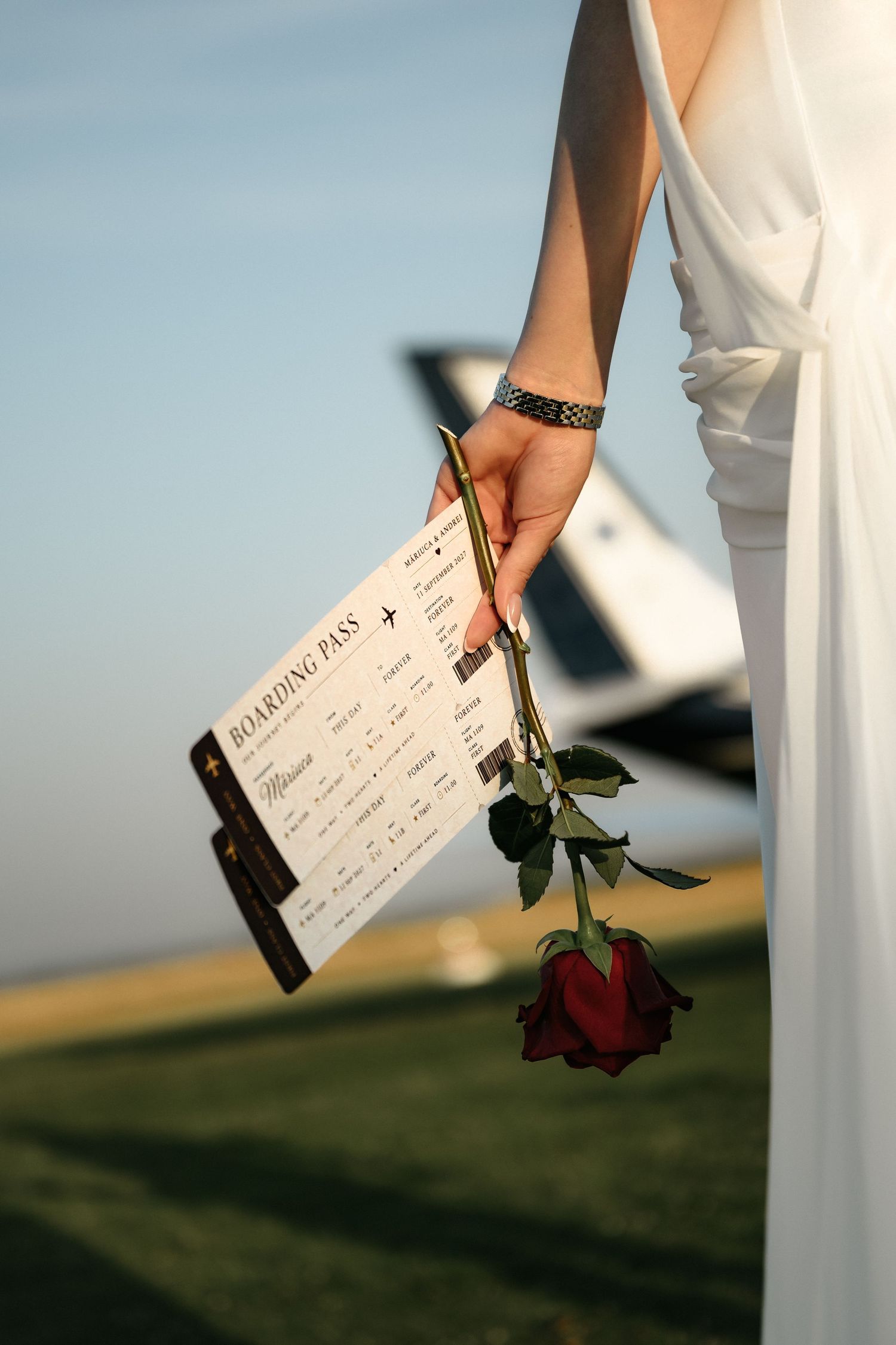 Bride in white dress holding a red rose and boarding pass near a private plane on a sunny day.