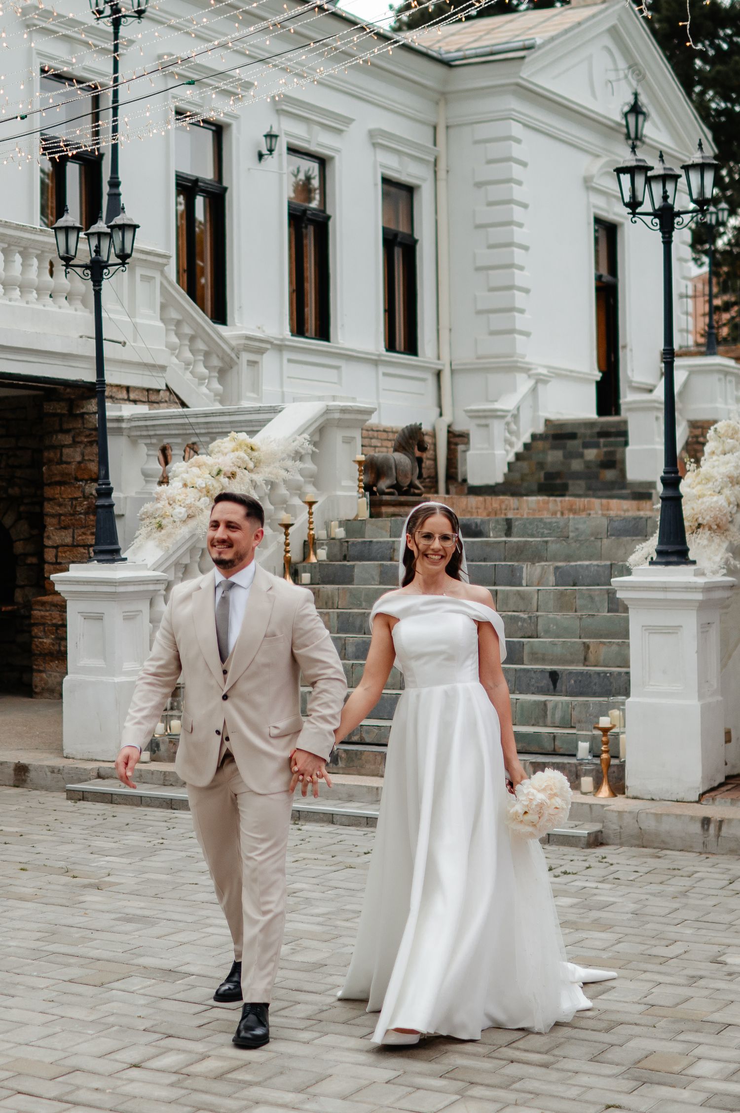 Smiling newlyweds walk hand in hand in front of white neoclassical wedding venue decorated with floral arrangements.
