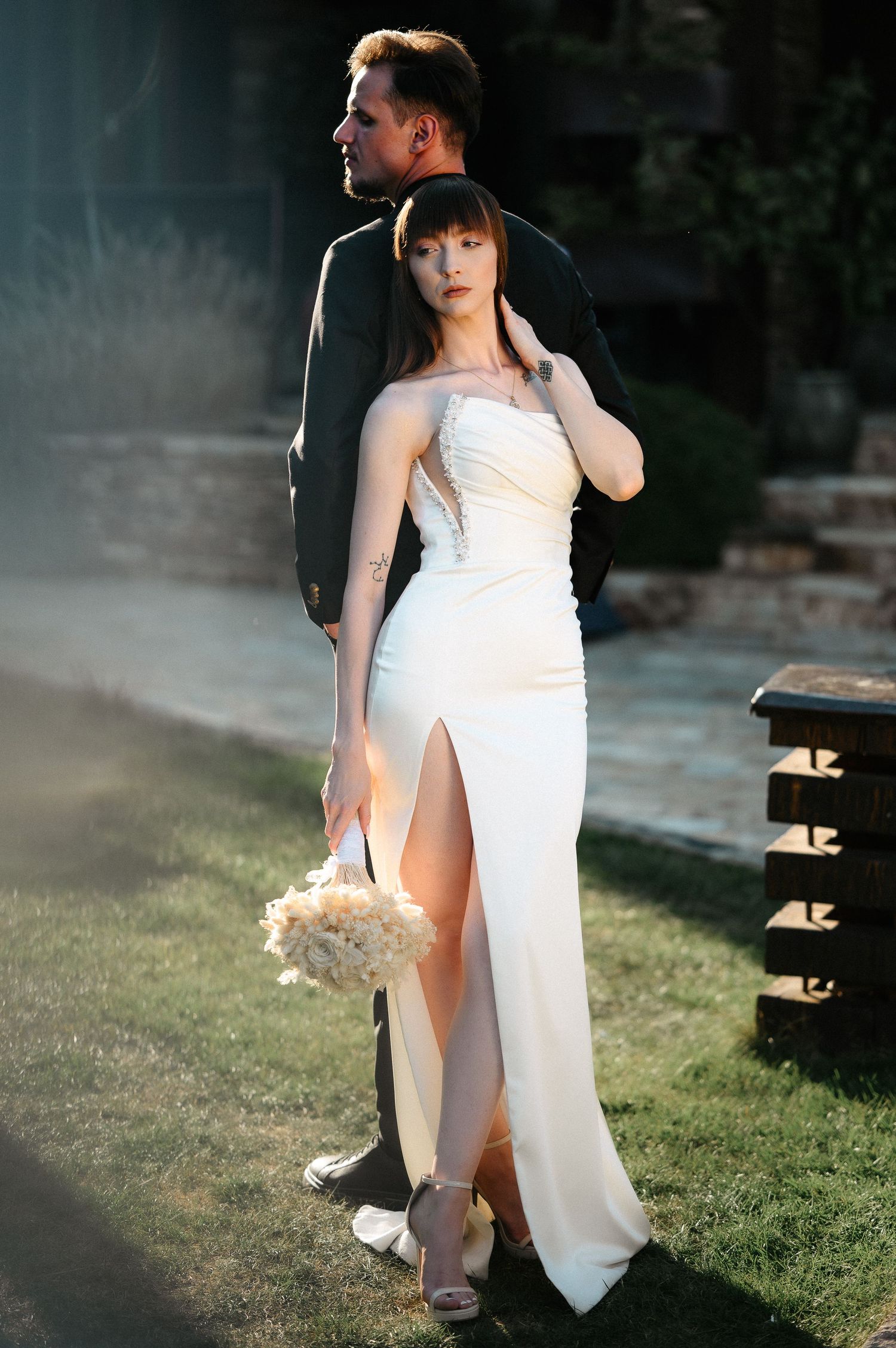 Bride in a strapless white slit gown with bouquet standing close to groom in black suit outdoors.