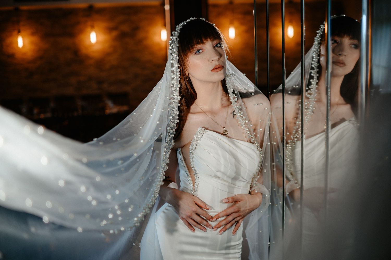 Elegant bride in strapless white gown with pearl-adorned veil poses by mirrors in warm light.
