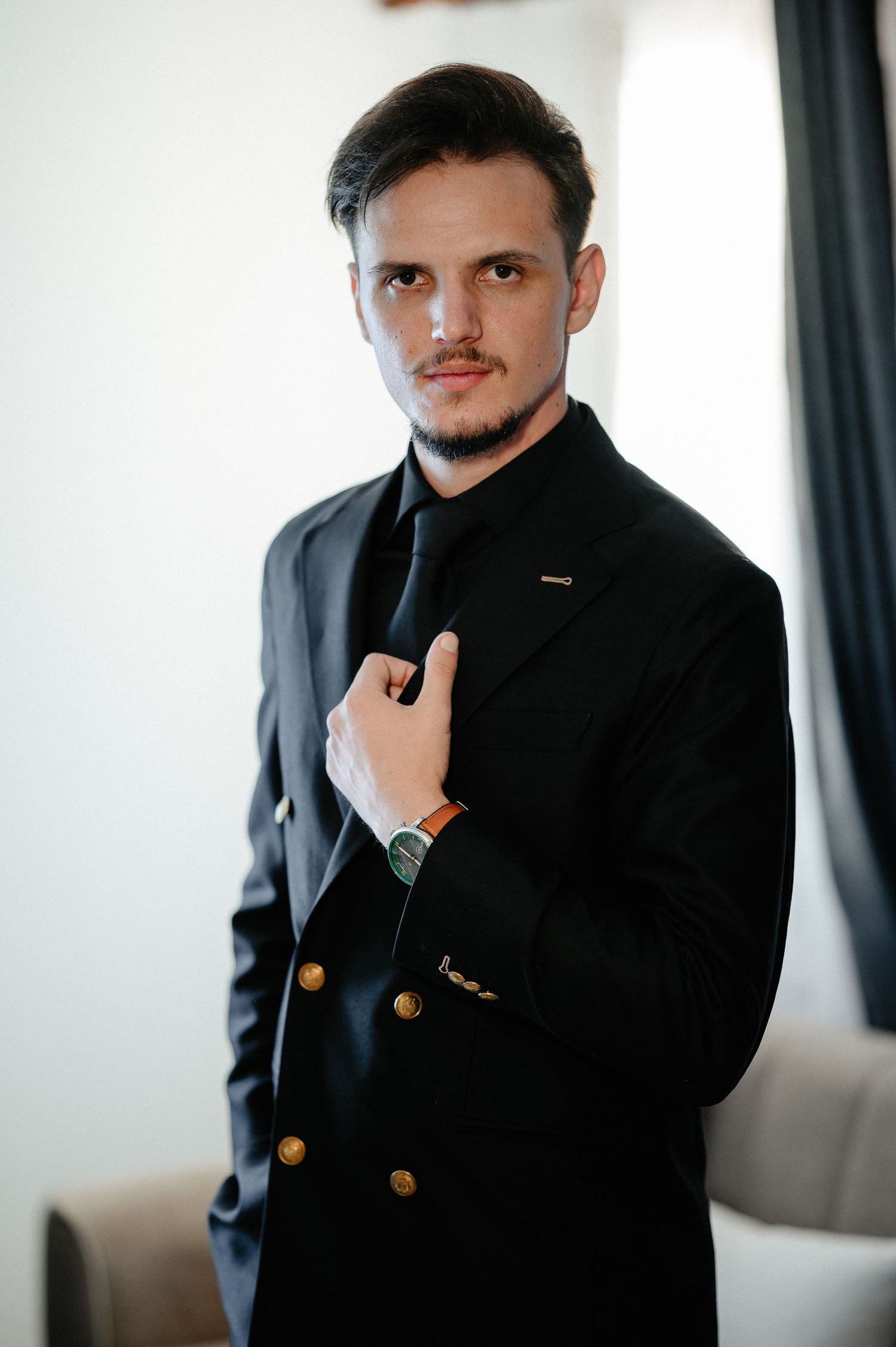 Groom in a black double-breasted suit with gold buttons adjusting his tie indoors.
