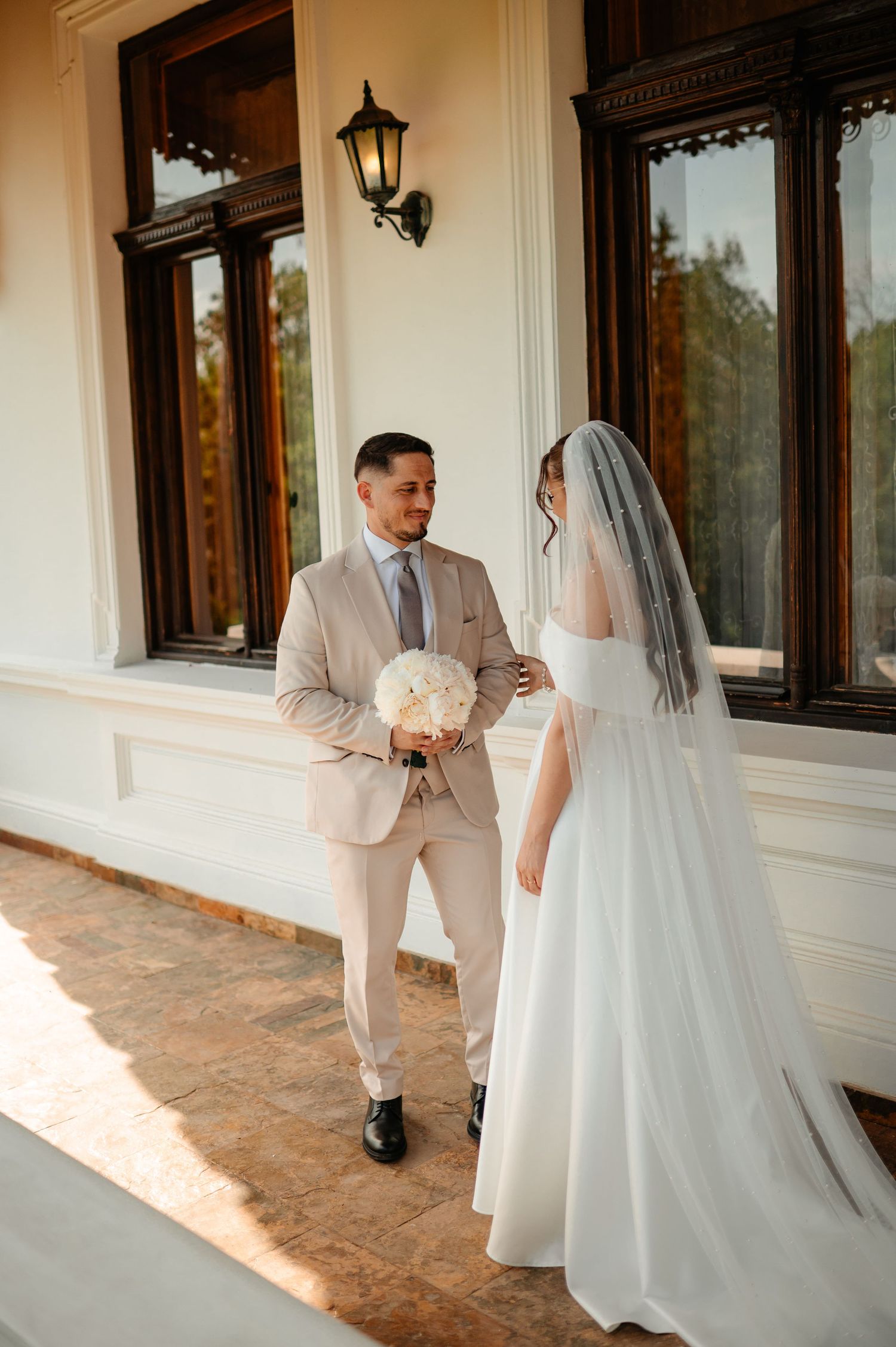 Groom in beige suit holds white bouquet facing bride in white off-shoulder gown with veil on elegant venue porch.